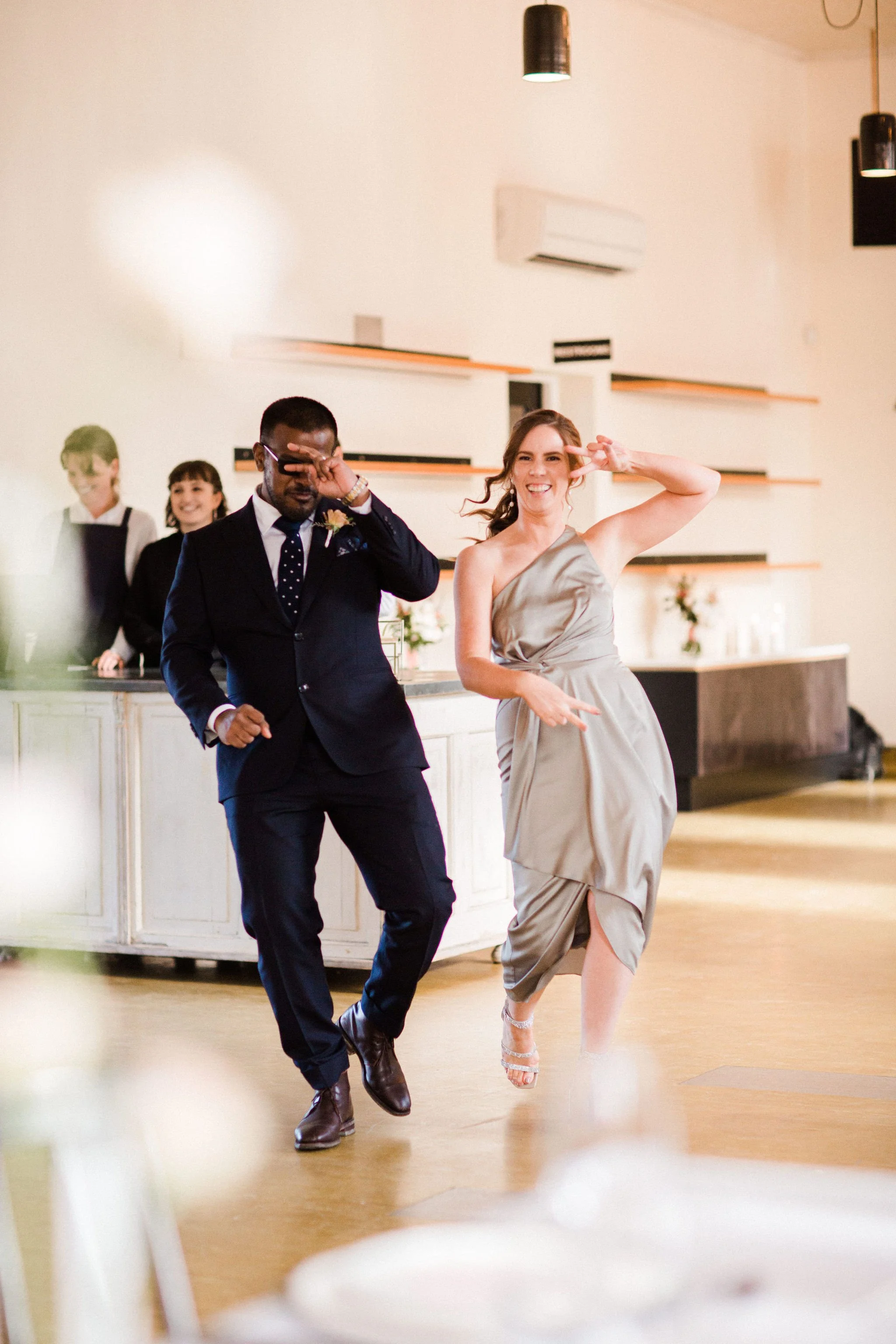 Two people dancing at a wedding reception, with three people in the background behind a bar. The woman is smiling and making a peace sign, while the man is dressed in a suit.