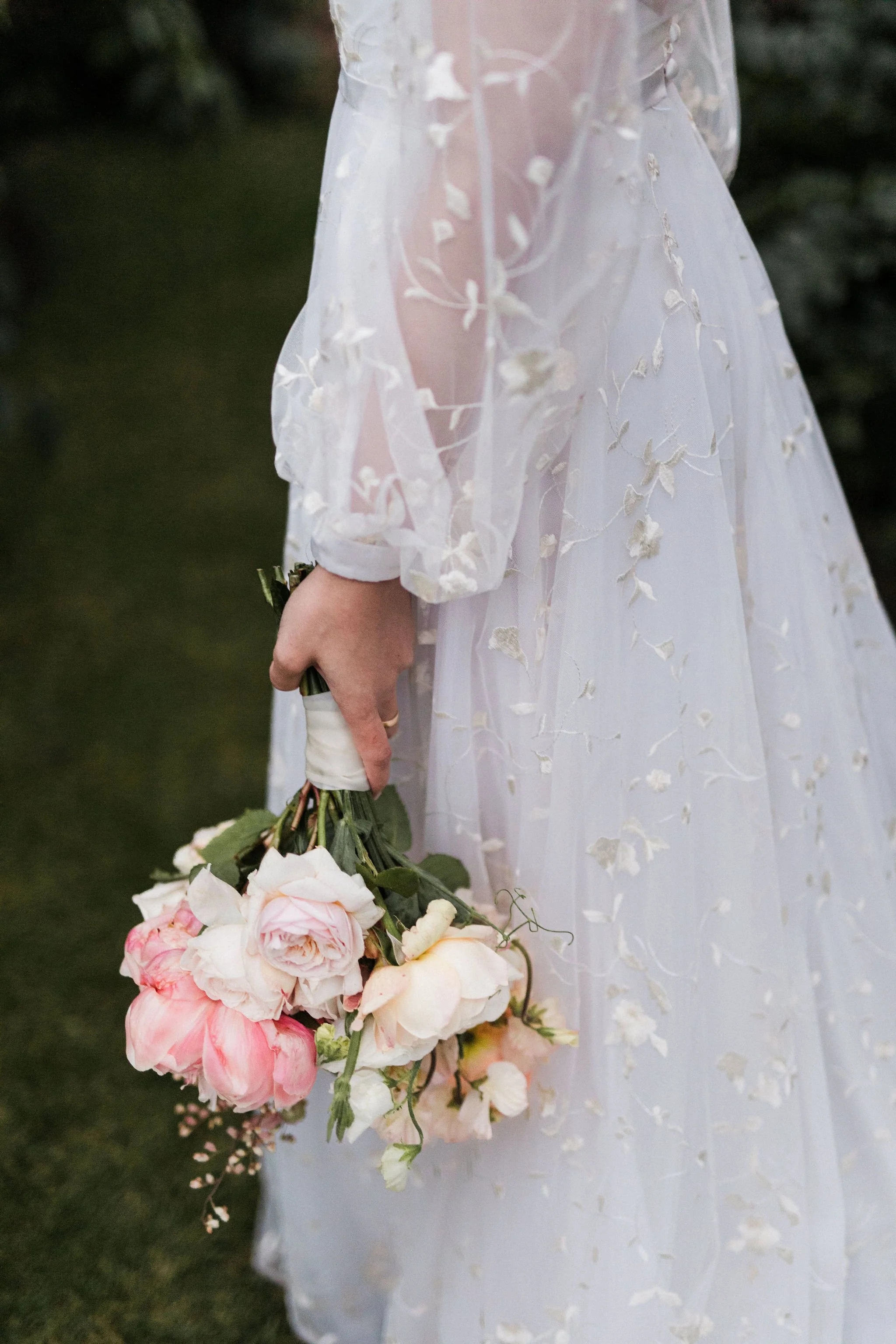 A person in a white, embroidered, sheer wedding dress holding a bouquet of pink and white roses.