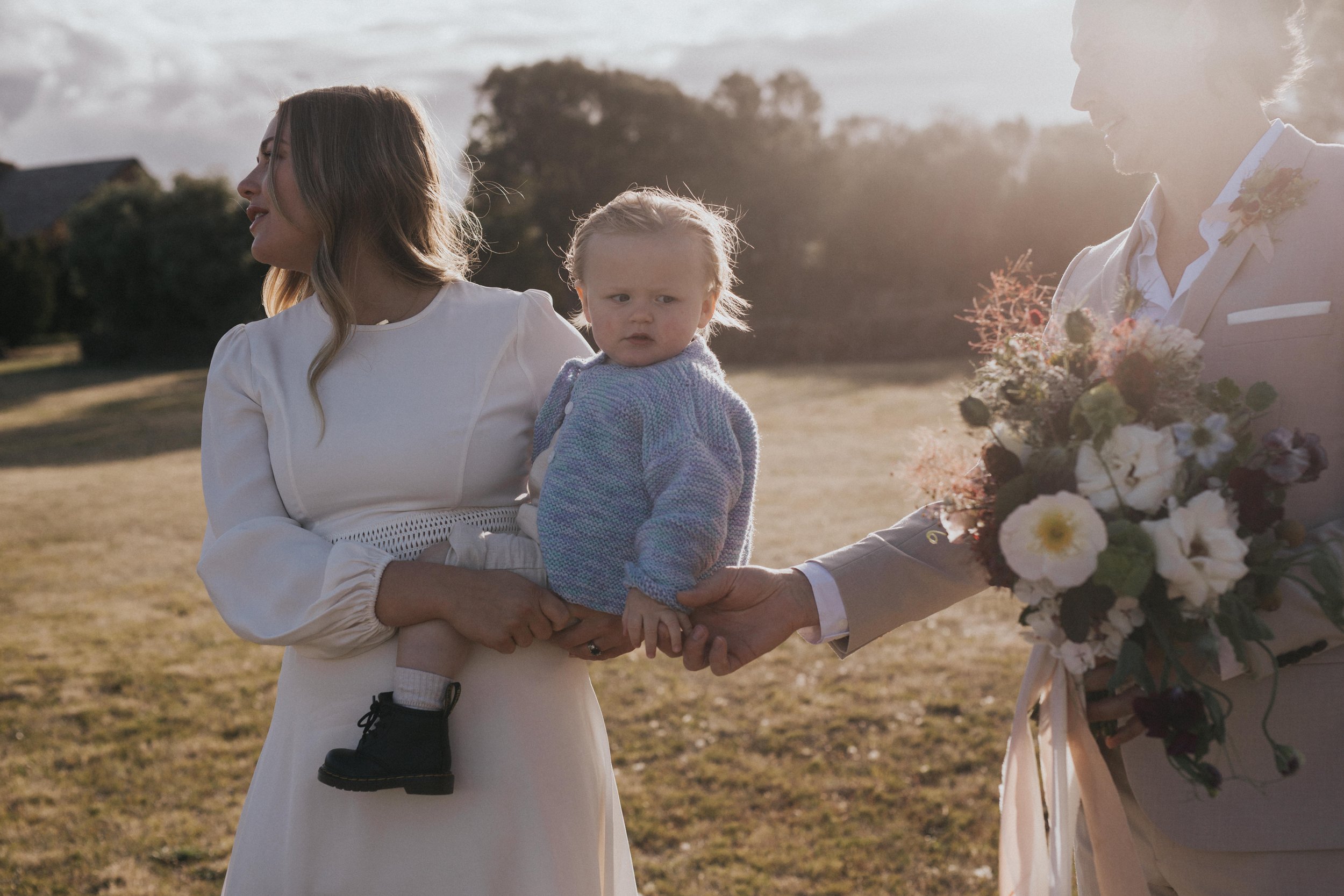 Children girl being held by woman, man holding flower bouquet, outdoor field, sunset lighting.