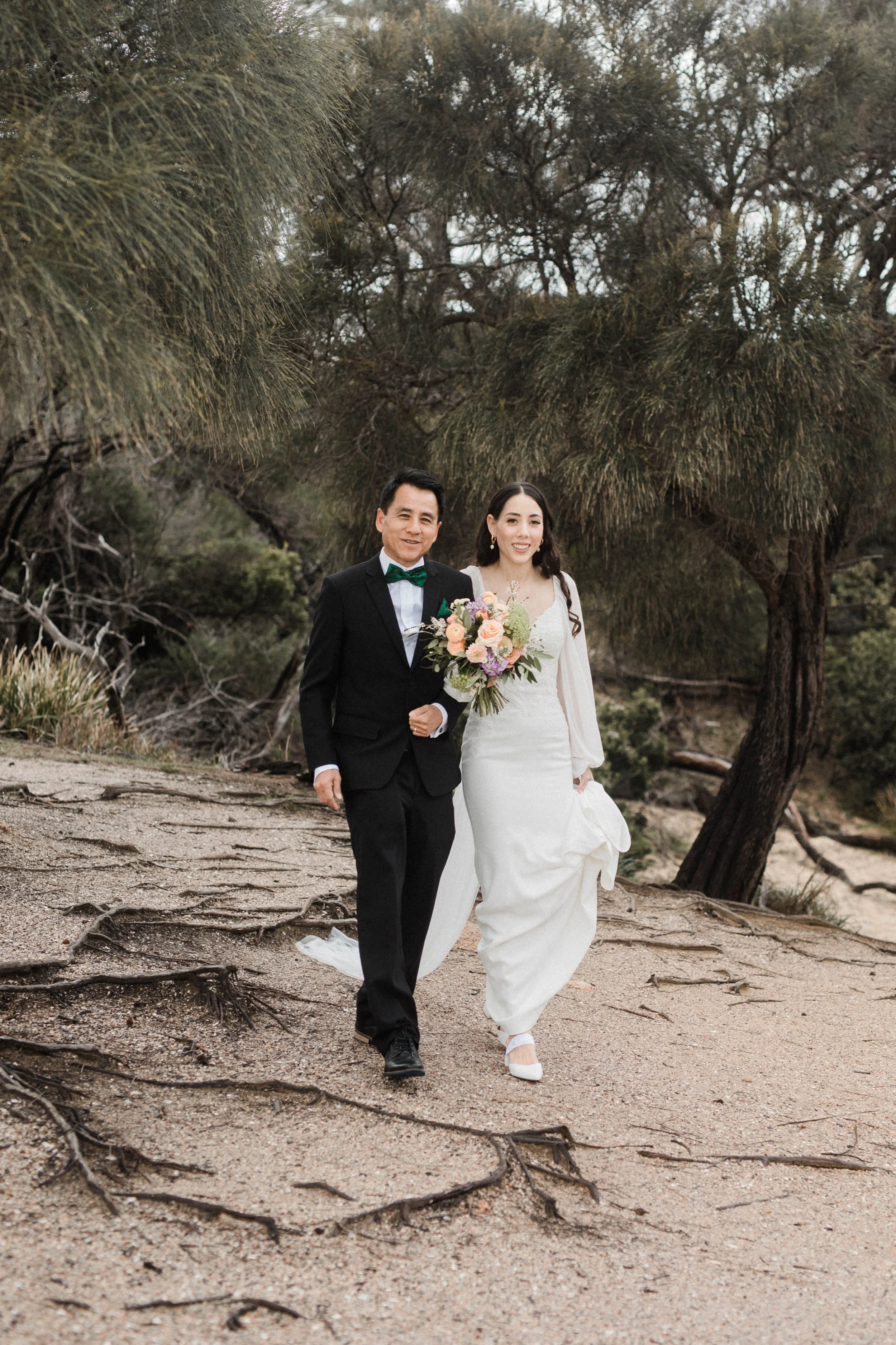 A bride in a white wedding dress holding a bouquet, walking with a groom in a black suit and green bow tie on a sandy trail surrounded by trees.