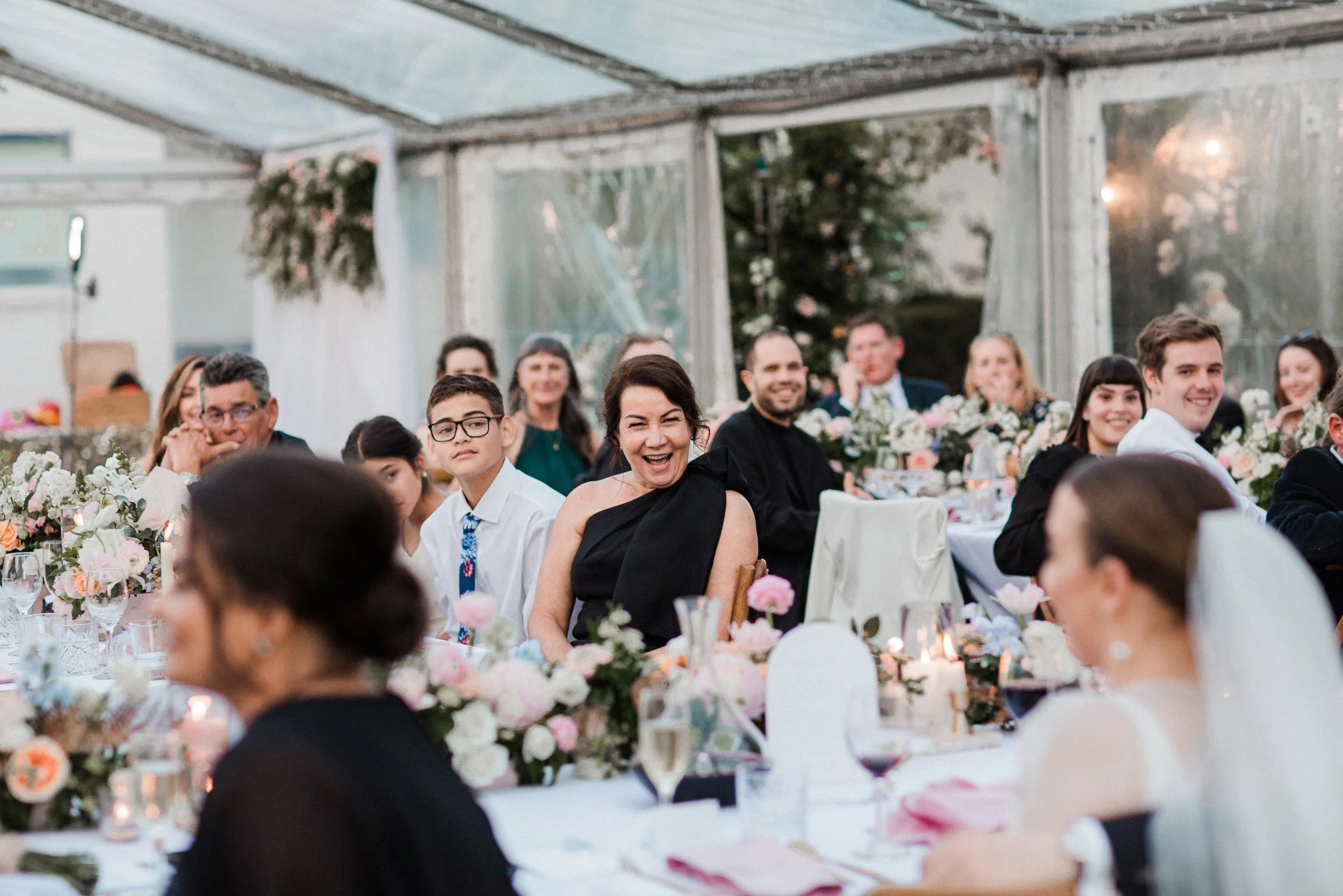 Guests sitting at a decorated wedding reception, smiling, under a clear tent with floral arrangements and candles.