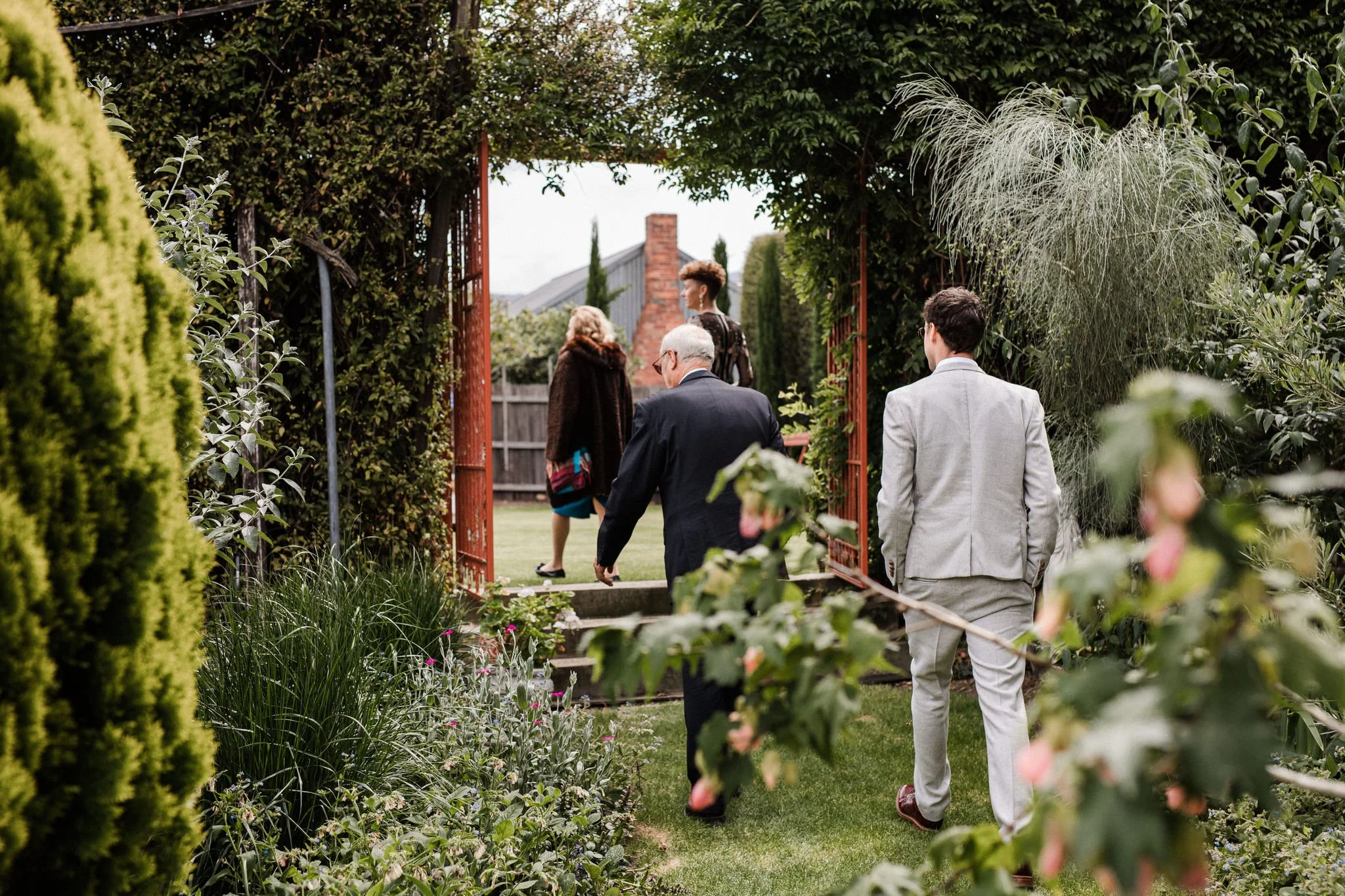 Several people walk through a garden gate into a backyard with a lawn and plants, with a brick chimney visible in the background.