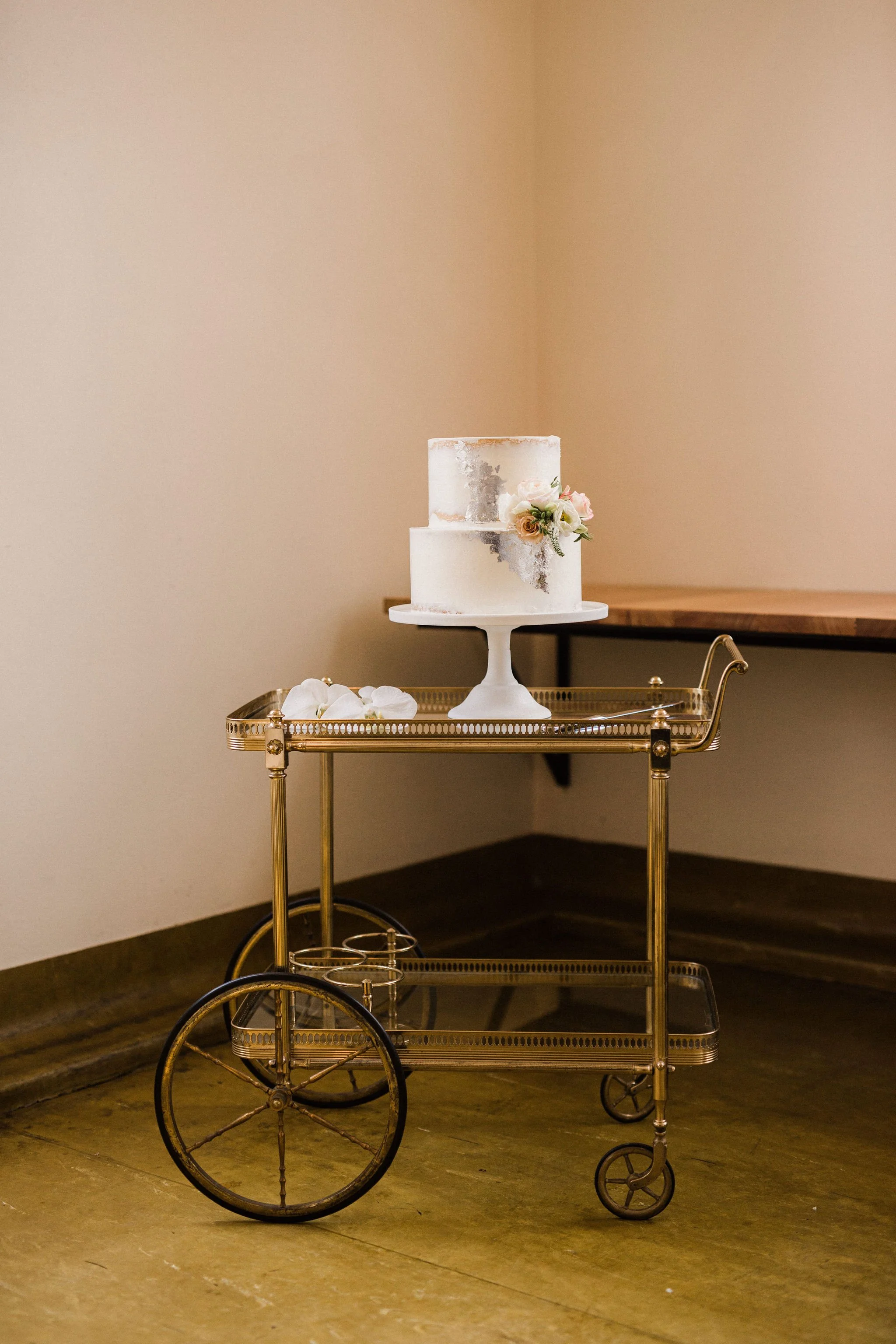 A two-tier white cake with a smooth finish and pink flowers as decoration on a white cake stand, placed on a vintage gold serving cart with wheels, on a wooden floor inside a room with a beige wall.