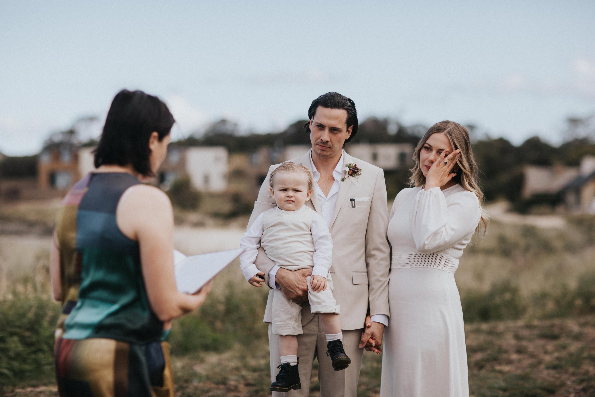 A man holding a young child, a woman in a white dress, and a person reading from a script outdoors during a wedding ceremony.