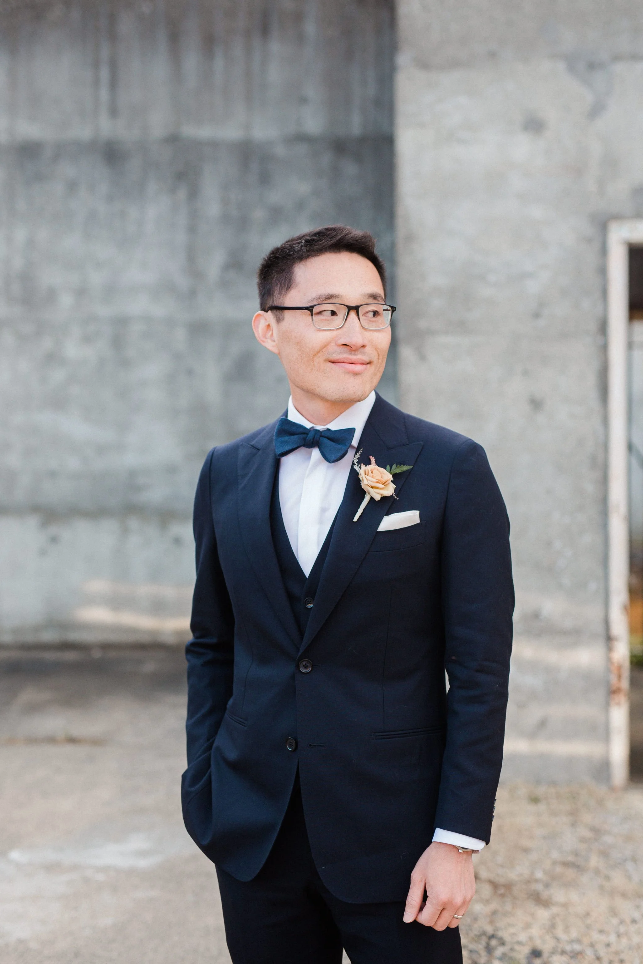 A man in a navy suit, white shirt, navy bow tie, and glasses, with a boutonniere, stands outdoors against a concrete wall, looking to the side.