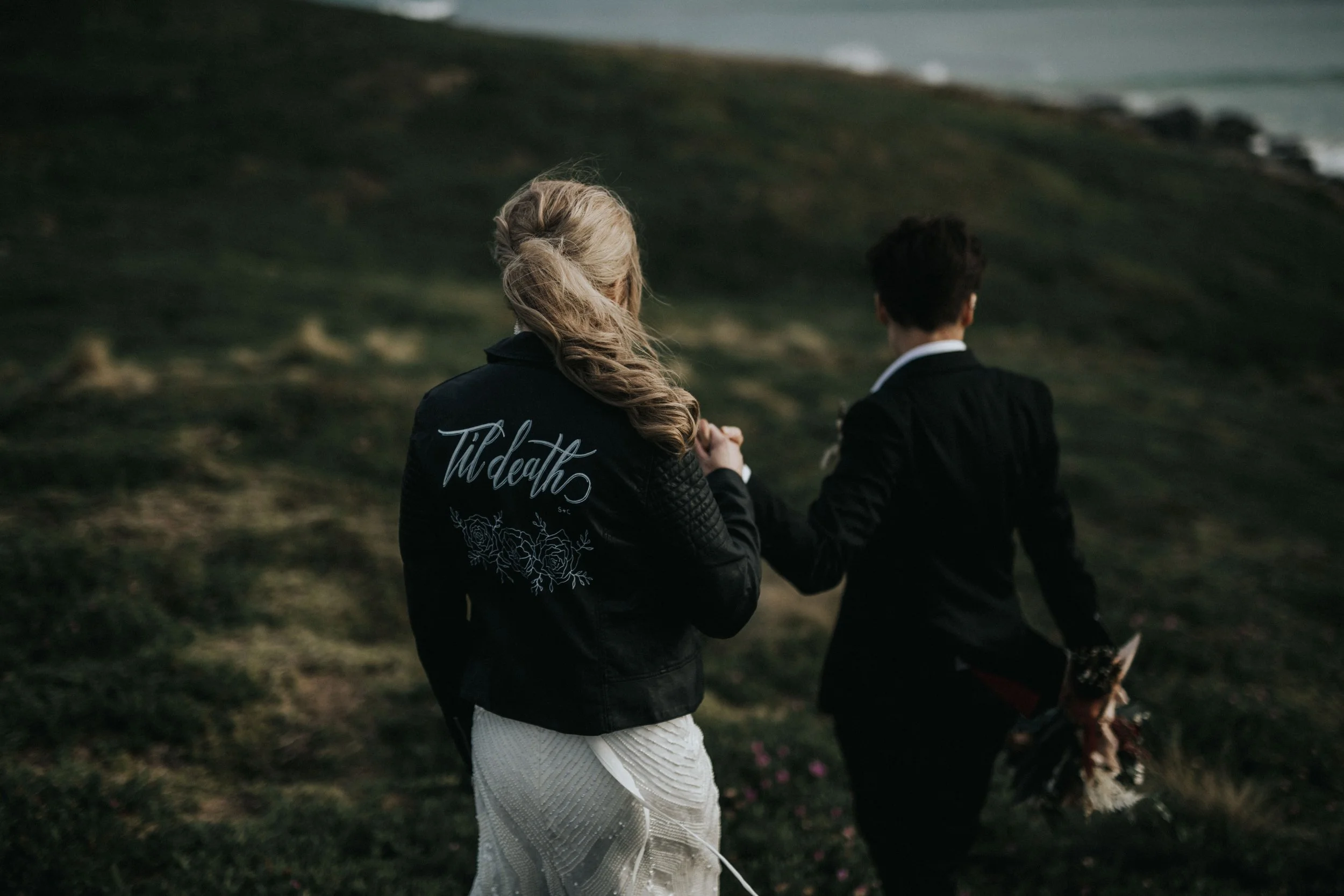A couple walking outdoors on a grassy hillside, with the woman wearing a black jacket with the words 'Til death' on the back and holding a bouquet of flowers in her hand.
