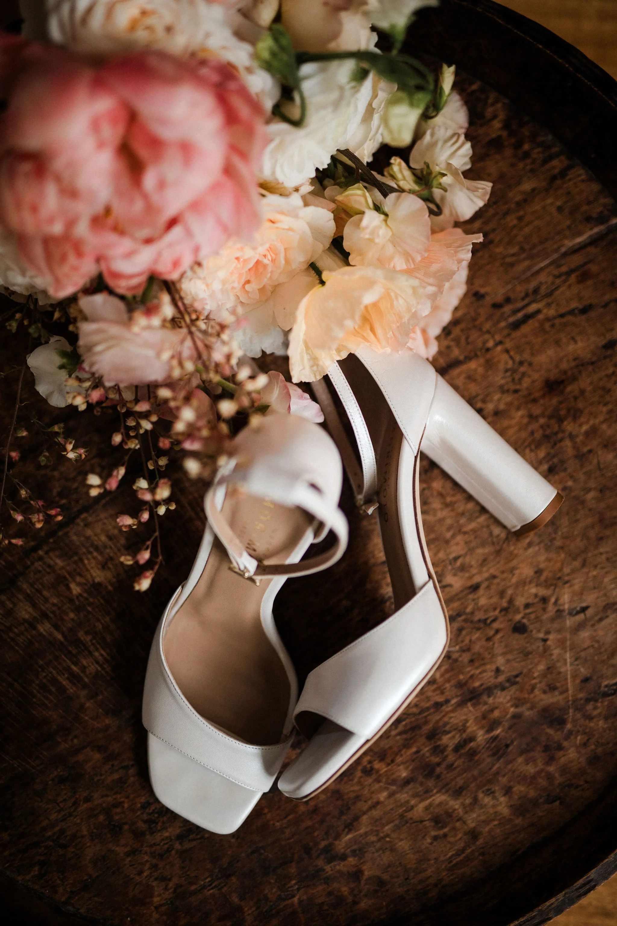 White high-heeled shoes with an open toe, placed on a dark wooden surface next to a bouquet of pink and white flowers.
