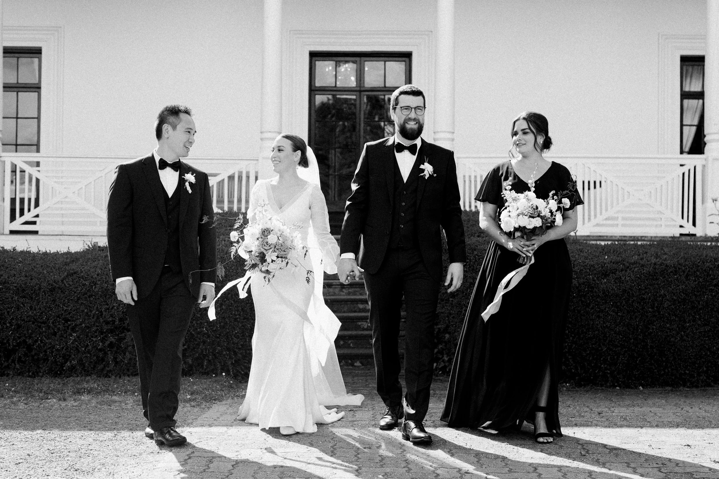 A black and white photo of a wedding party walking in front of a large house with stairs. The group includes a bride in a white wedding gown holding a bouquet, a groom in a suit holding the bride's hand, a man in a suit, and a woman in a black dress 