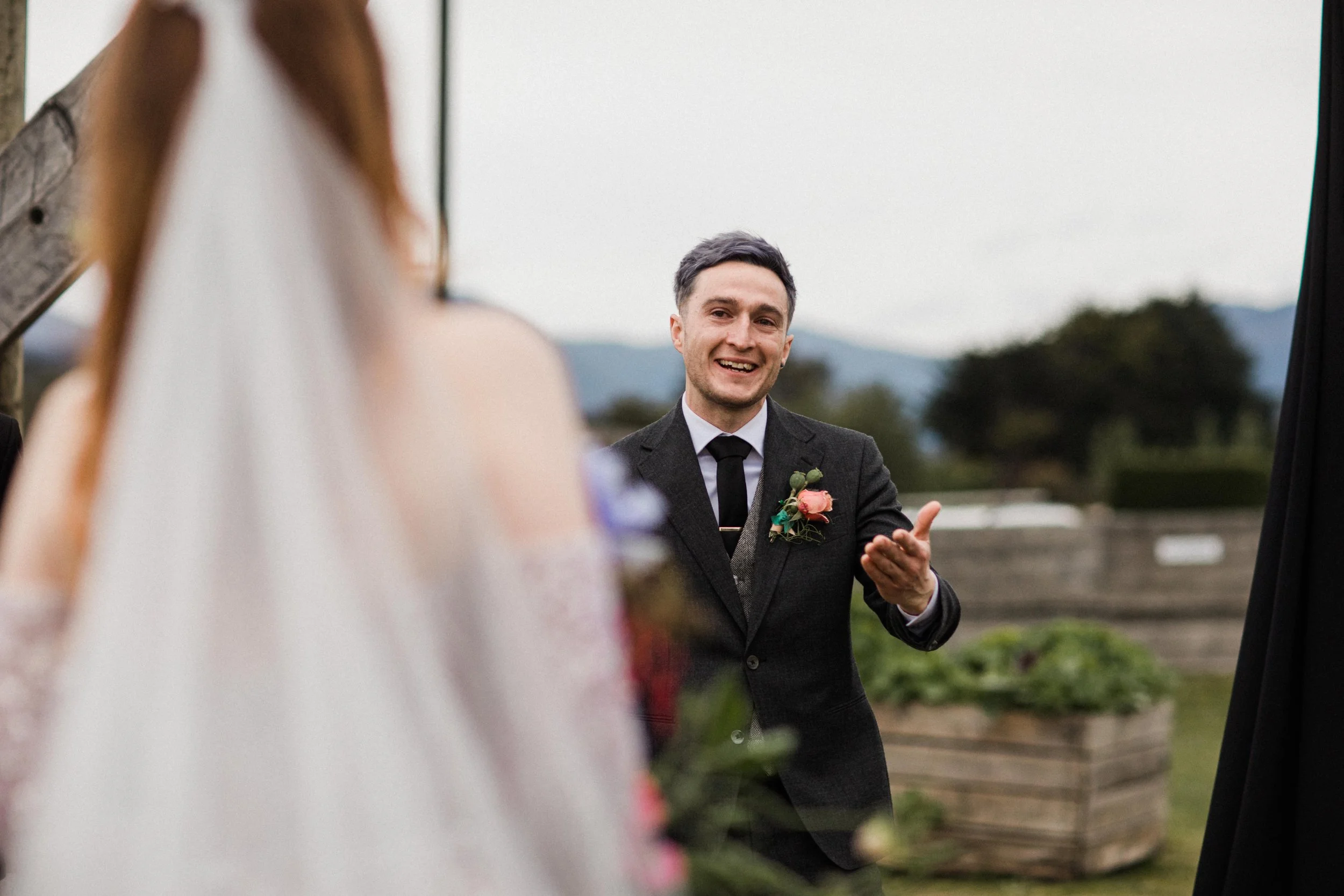A man in a dark suit with a pink rose boutonniere smiling and gesturing with his hand at an outdoor wedding ceremony, with a person in a white dress partially visible in the foreground.