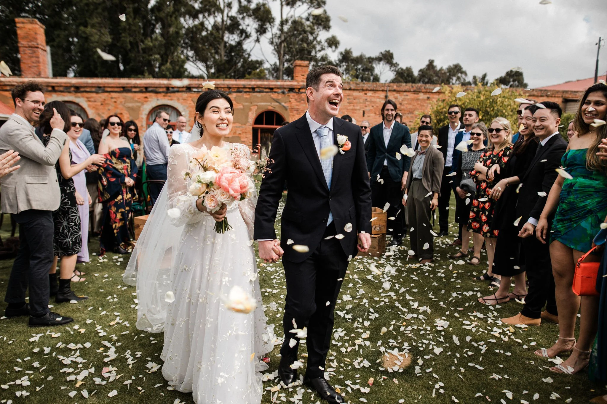 Happy bride and groom walking hand in hand through a confetti explosion at their outdoor wedding, surrounded by guests celebrating.