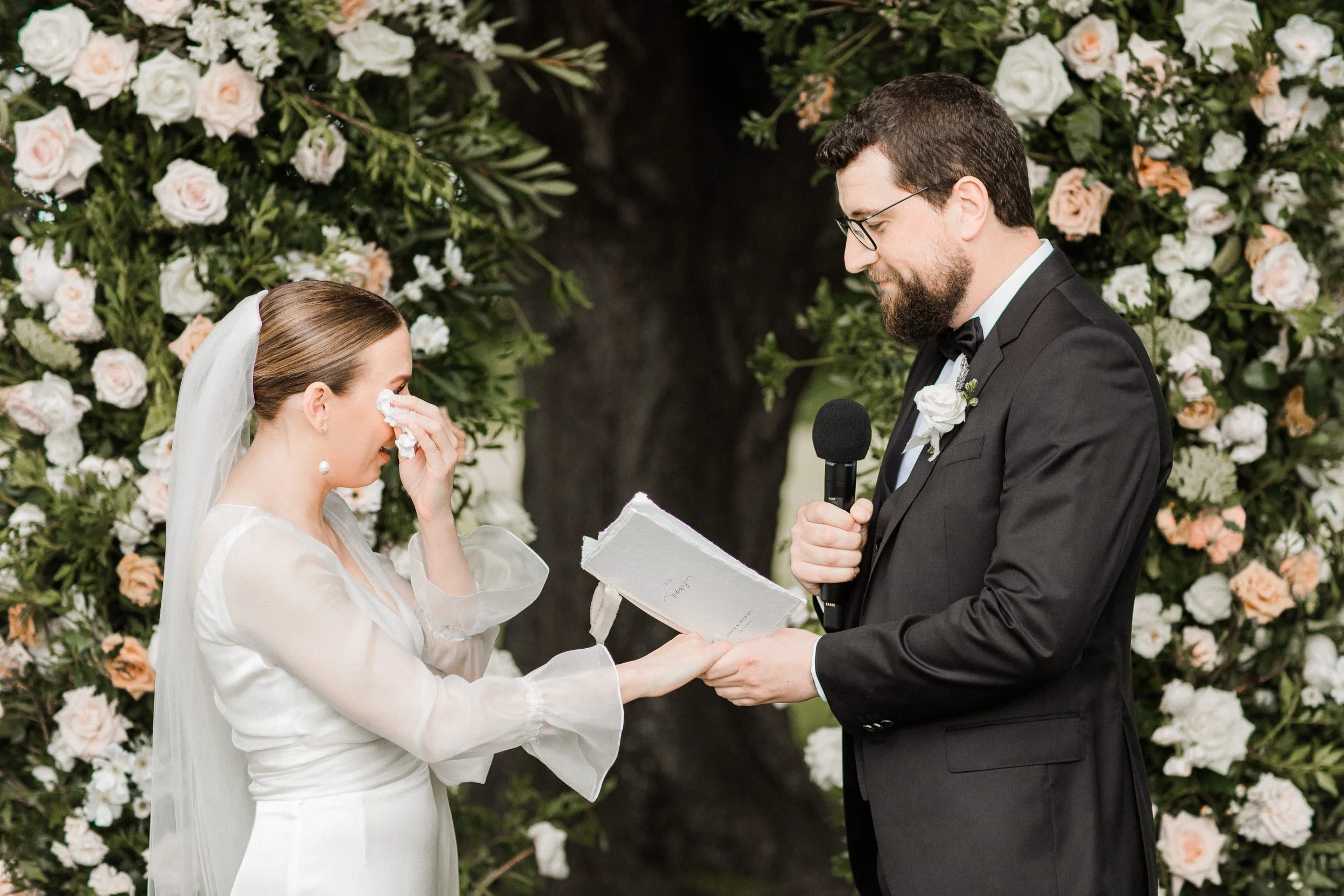 A bride wiping tears during her wedding ceremony while a groom reads vows, standing in front of a floral backdrop.