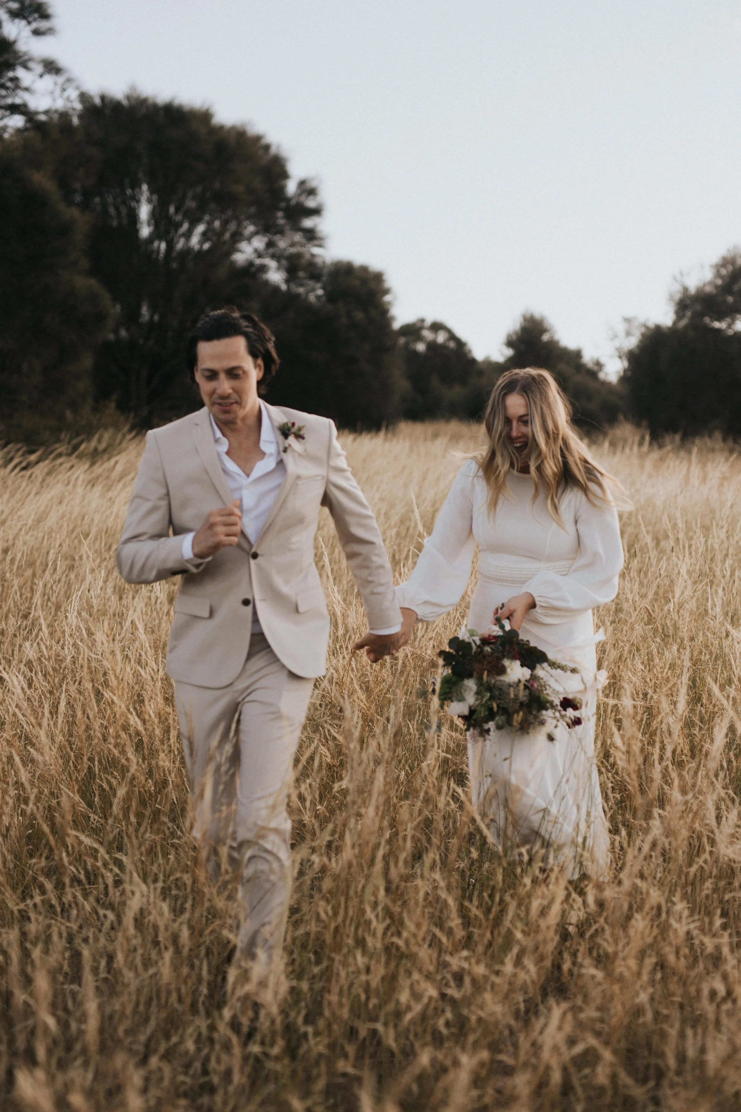 A couple dressed in wedding attire, holding hands and walking through a grassy field with trees in the background.