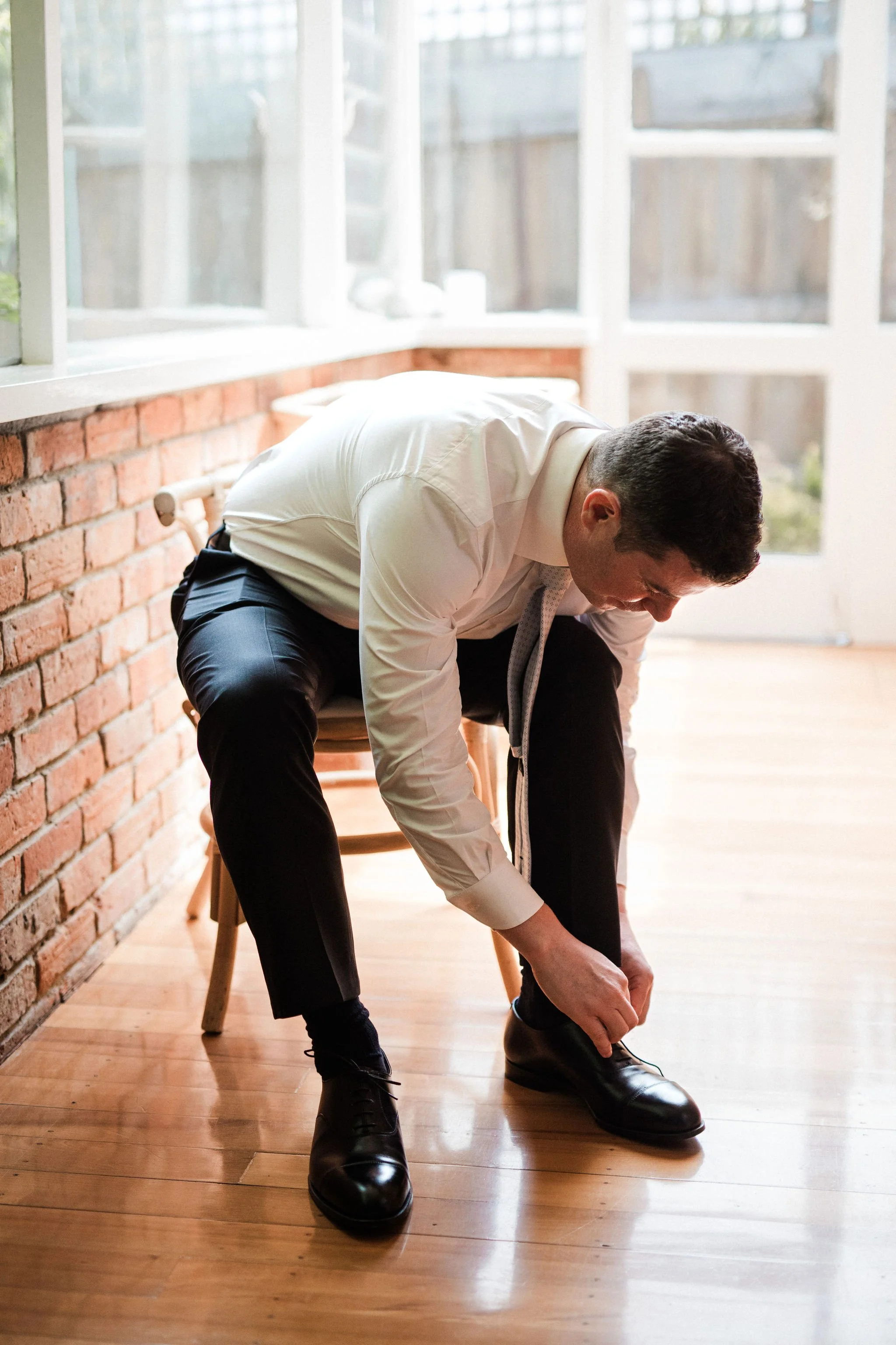 A man in dress clothes is sitting on a wooden chair, leaning forward to tie his black dress shoes.