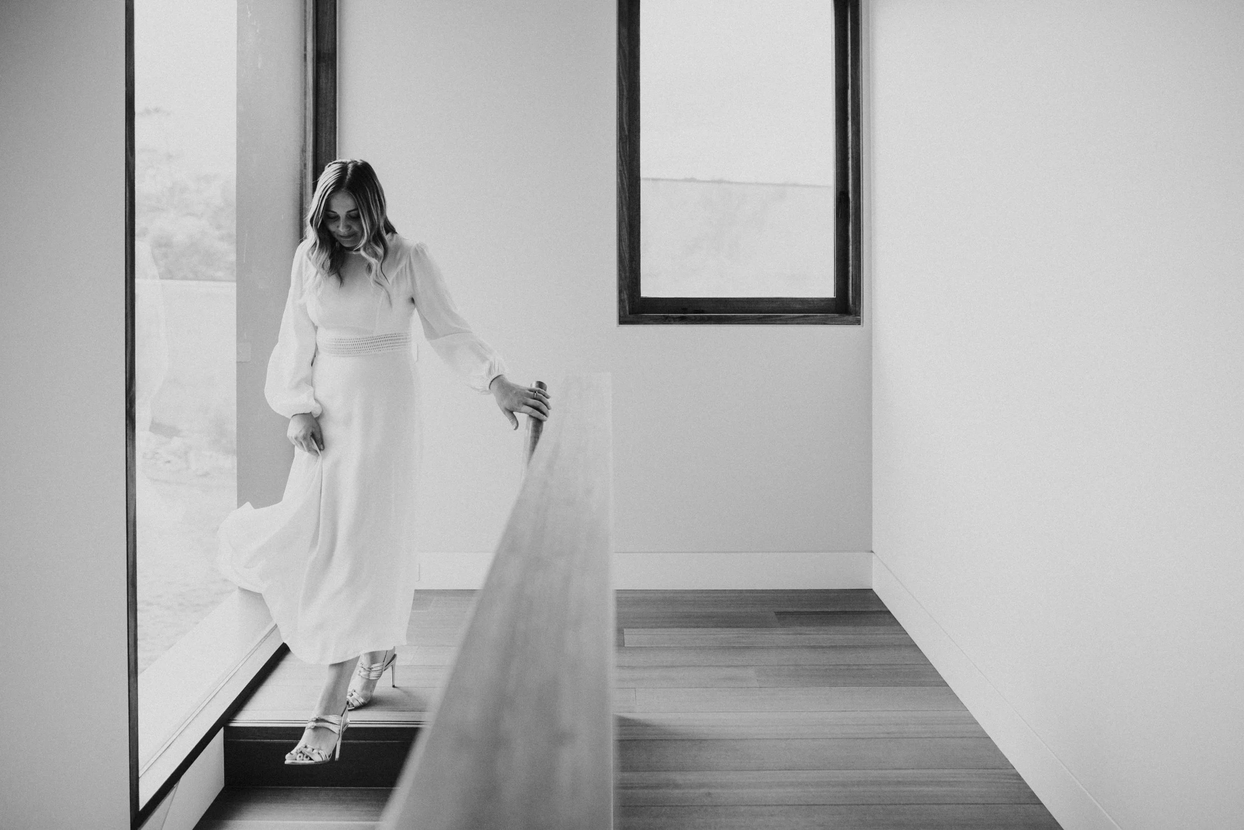 A woman in a white dress walking down stairs next to a large window, black and white photo.