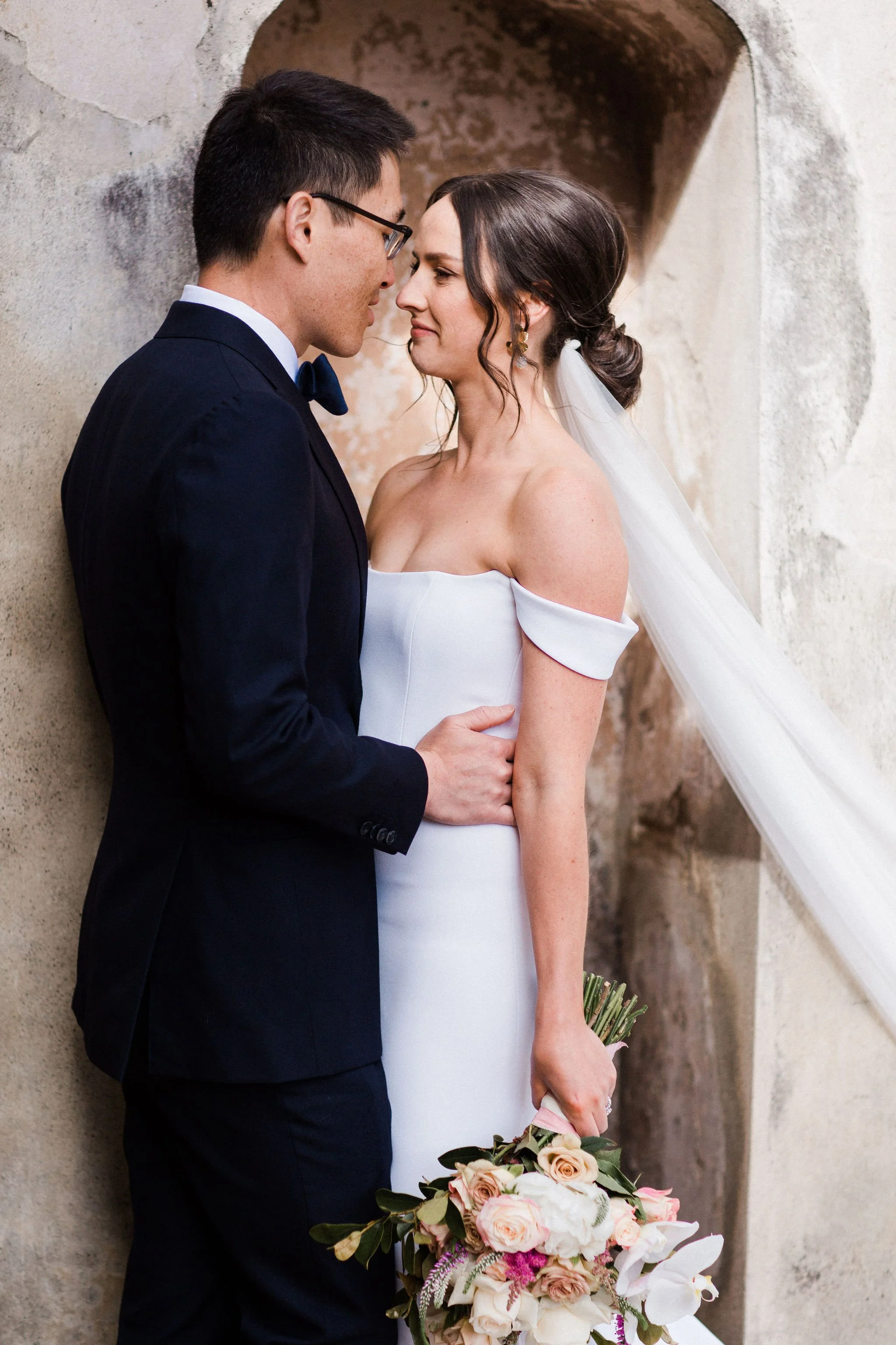A bride and groom standing close together, touching foreheads, with a rustic wall in the background. The bride is holding a bouquet of flowers.