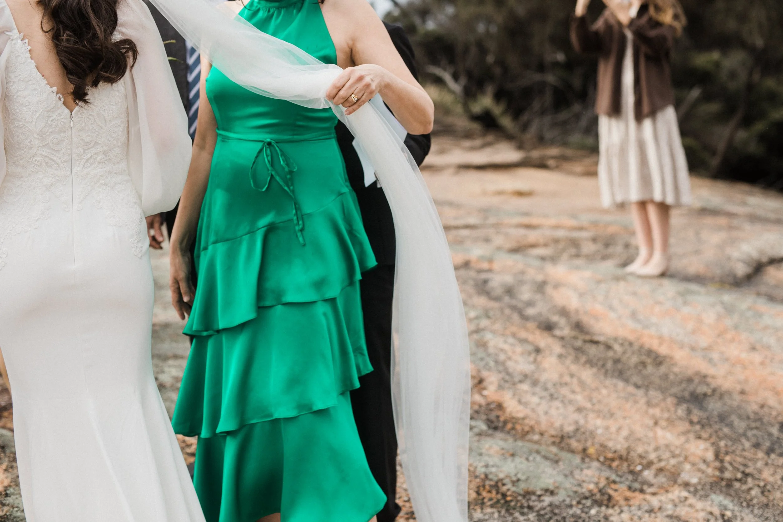 People standing outdoors on rocky ground, with a woman in a green layered dress in the center, a woman in a white dress to the left, and another woman in a striped skirt and brown jacket in the background.