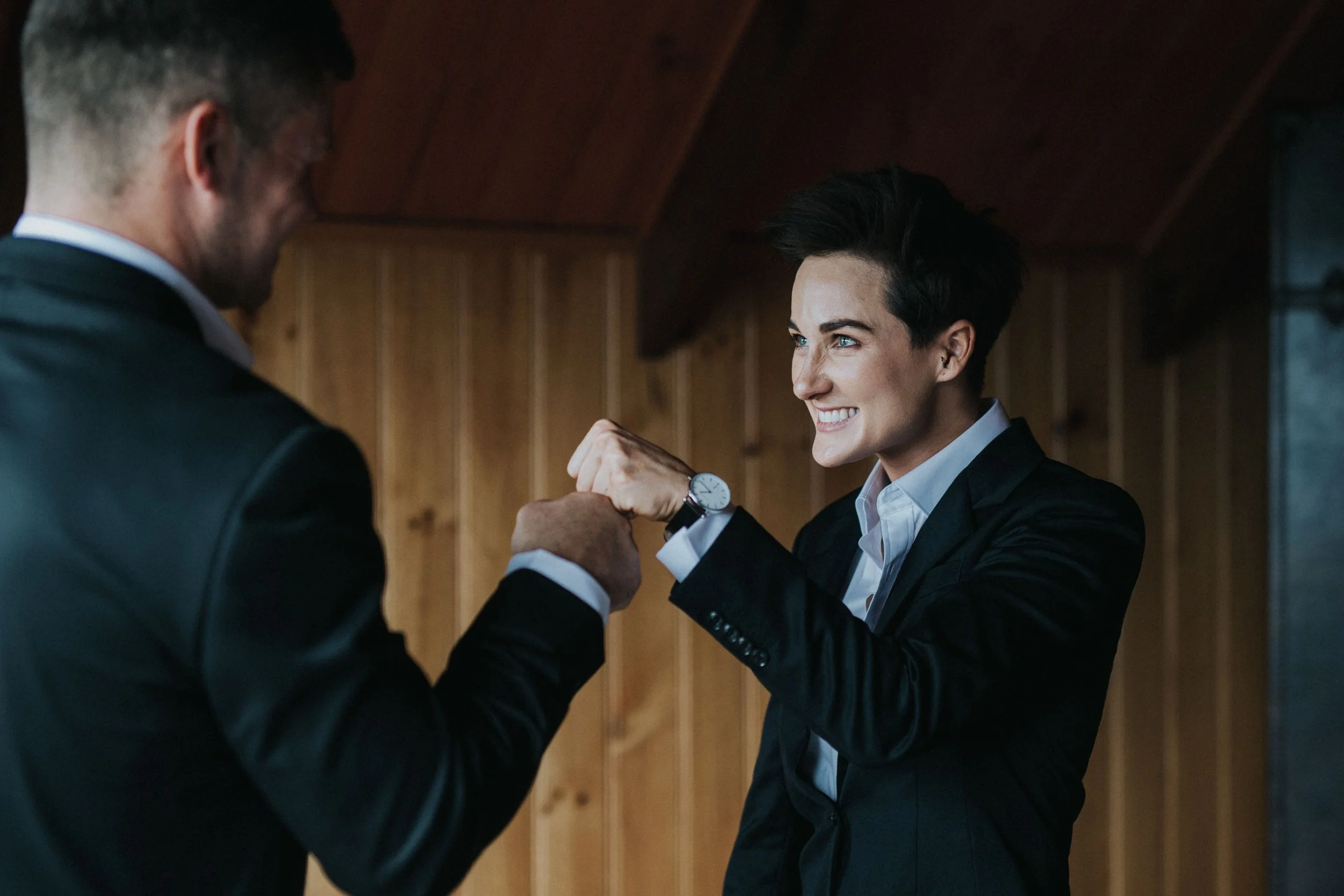 Two people, a woman and a man, smiling and bumping fists in a friendly, professional setting with wooden walls.