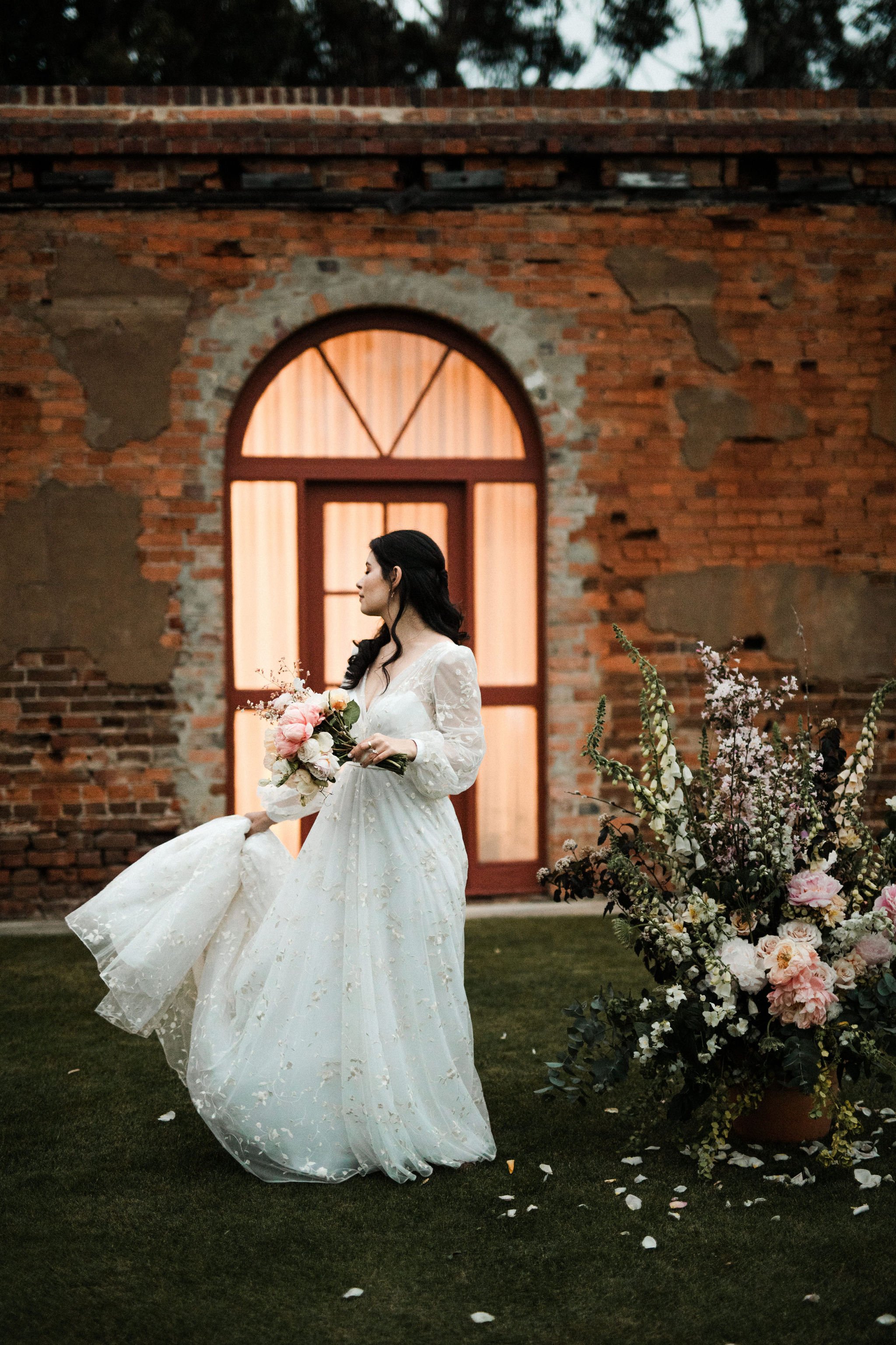 A bride holding a bouquet of pink and white flowers, wearing a white, long-sleeved wedding dress with lace details, standing outdoors on grass near a brick wall with a large arched window behind her and a big floral arrangement nearby.