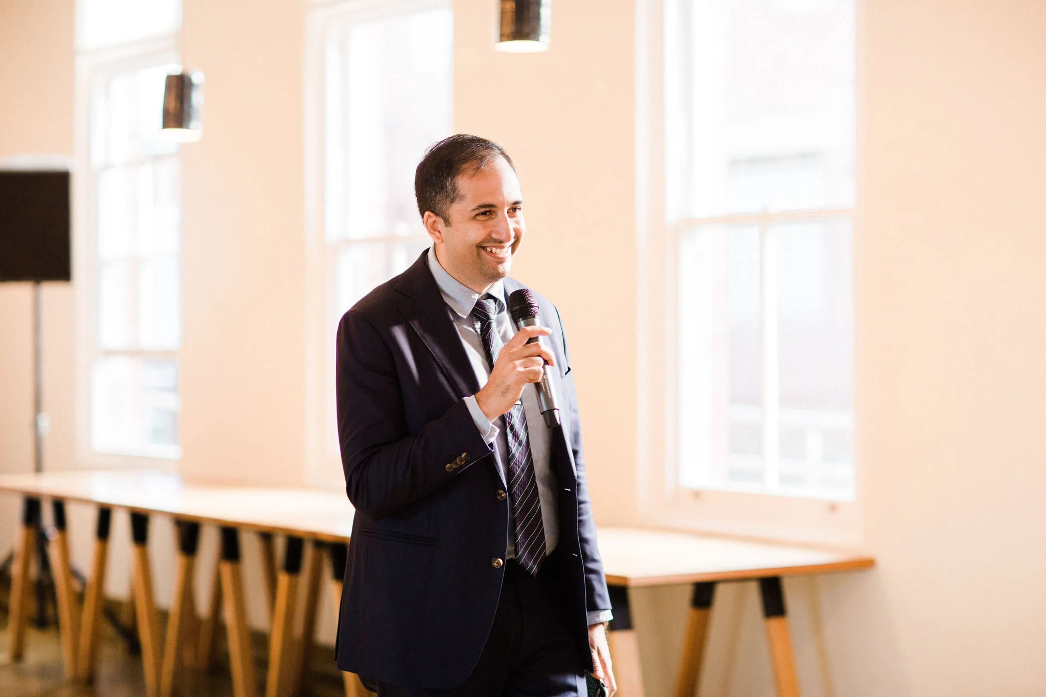 A man in a suit holding a microphone and smiling in a well-lit room with large windows and a long table.