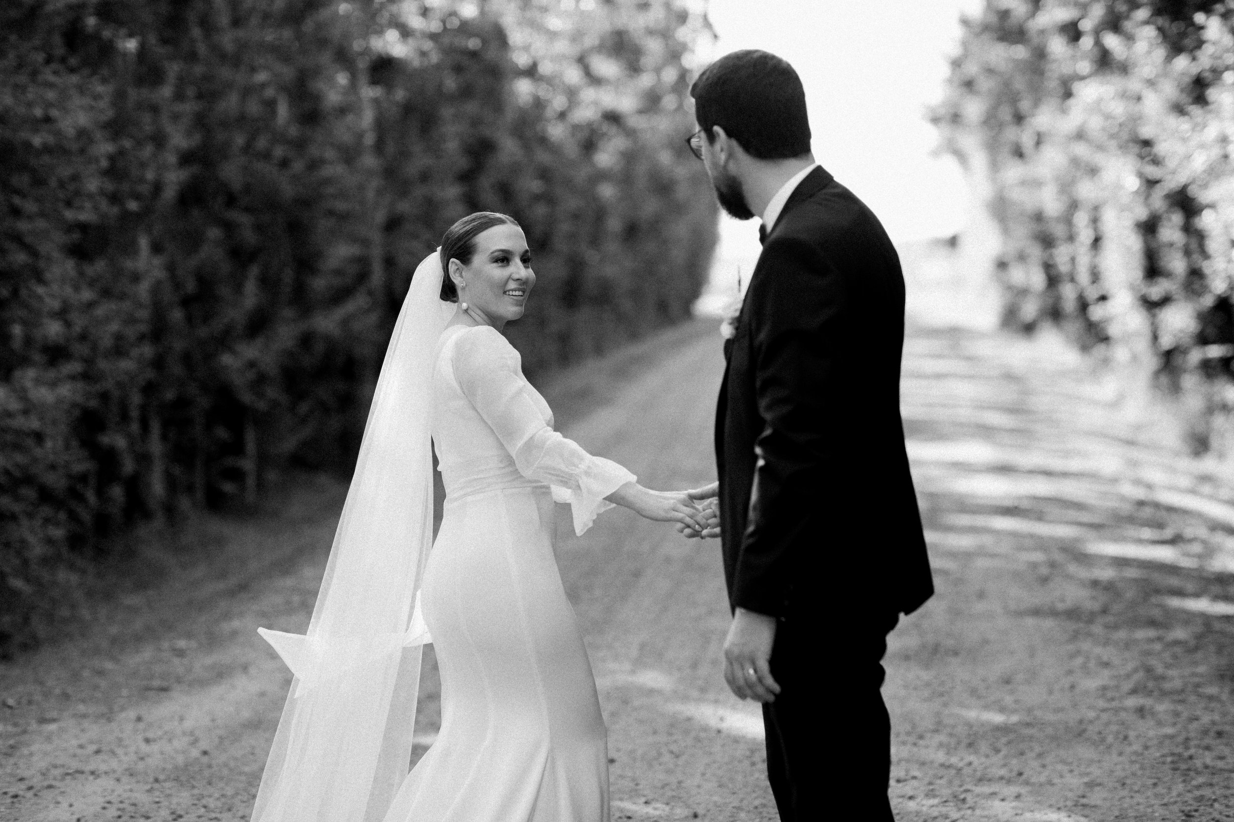 A bride and groom holding hands and smiling at each other outdoors.