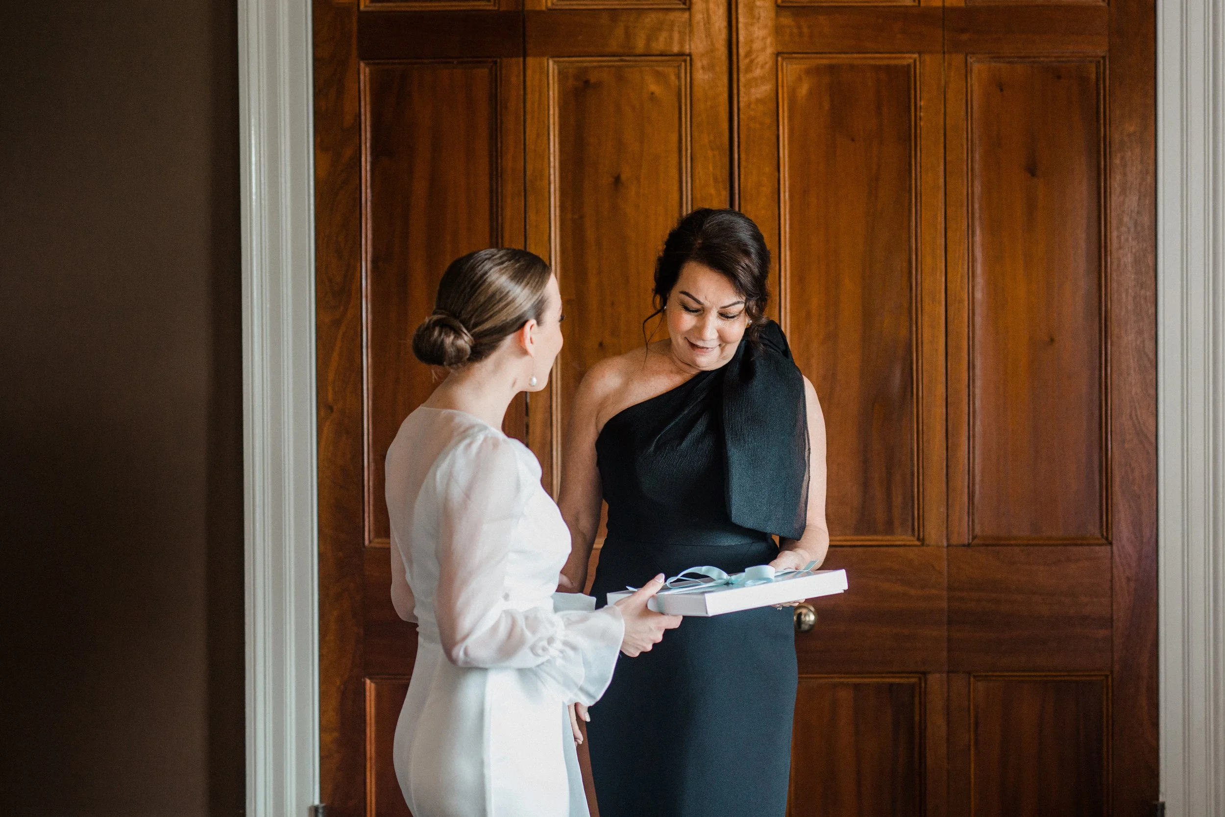 Two women standing in front of a wood paneled door, one wearing a white outfit with her hair in a bun, the other in a black dress with dark hair, holding a gift box with a blue ribbon, smiling as they converse.