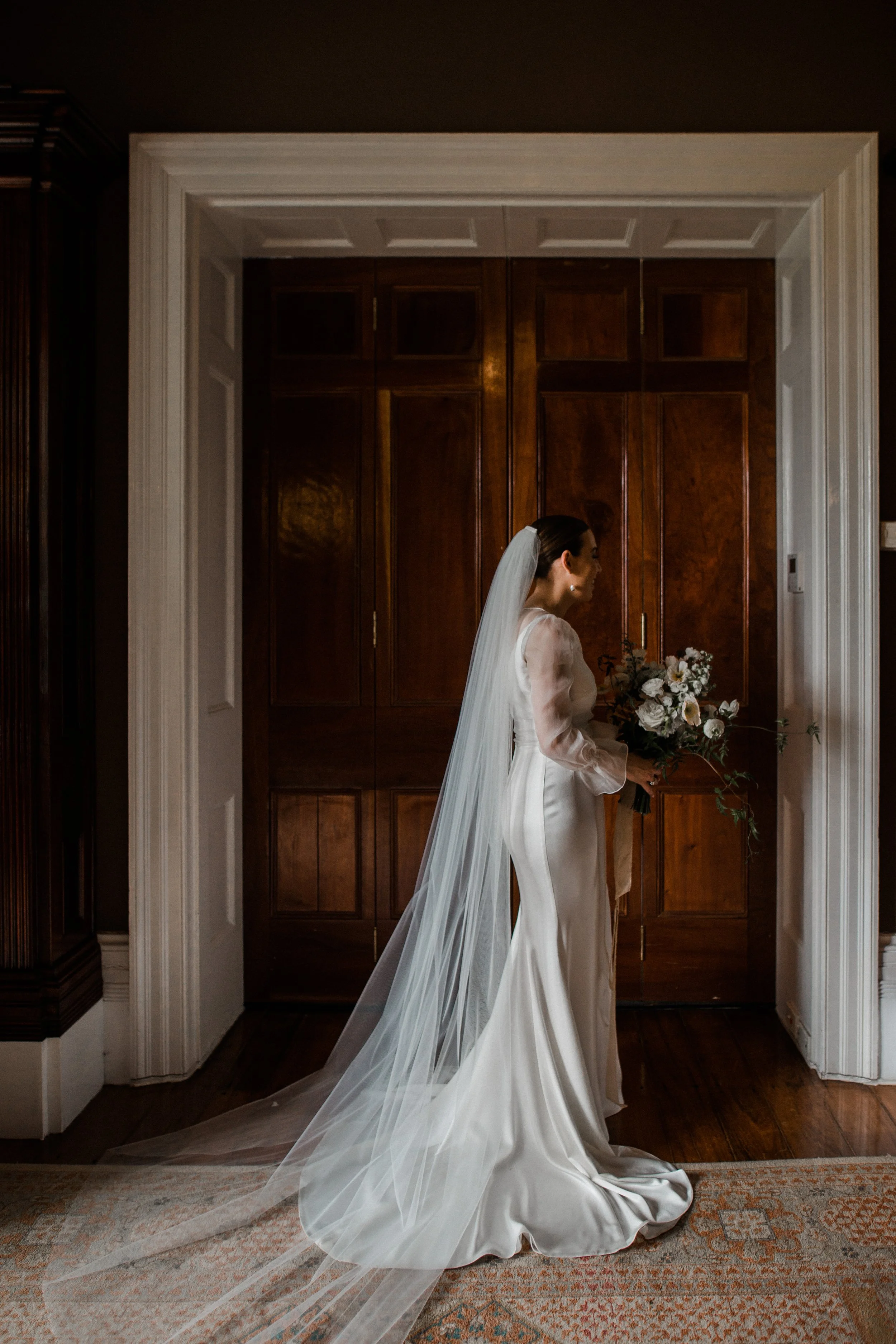 A bride in a long white satin wedding gown with a veil, holding a bouquet of flowers, standing in front of wooden doors in a dimly lit room.