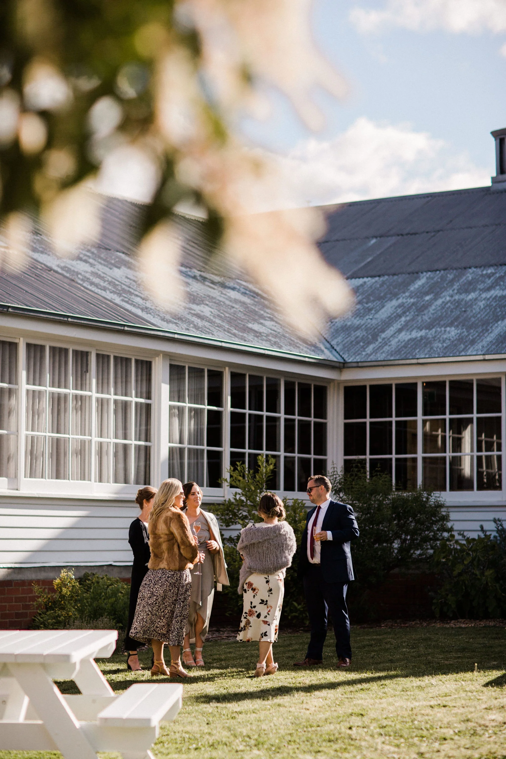 Group of people socializing outside a house during daytime, dressed in semi-formal attire, with one woman holding a glass of champagne, and a man in a suit. The background features a house with large windows and a leafless tree branch in the foregrou