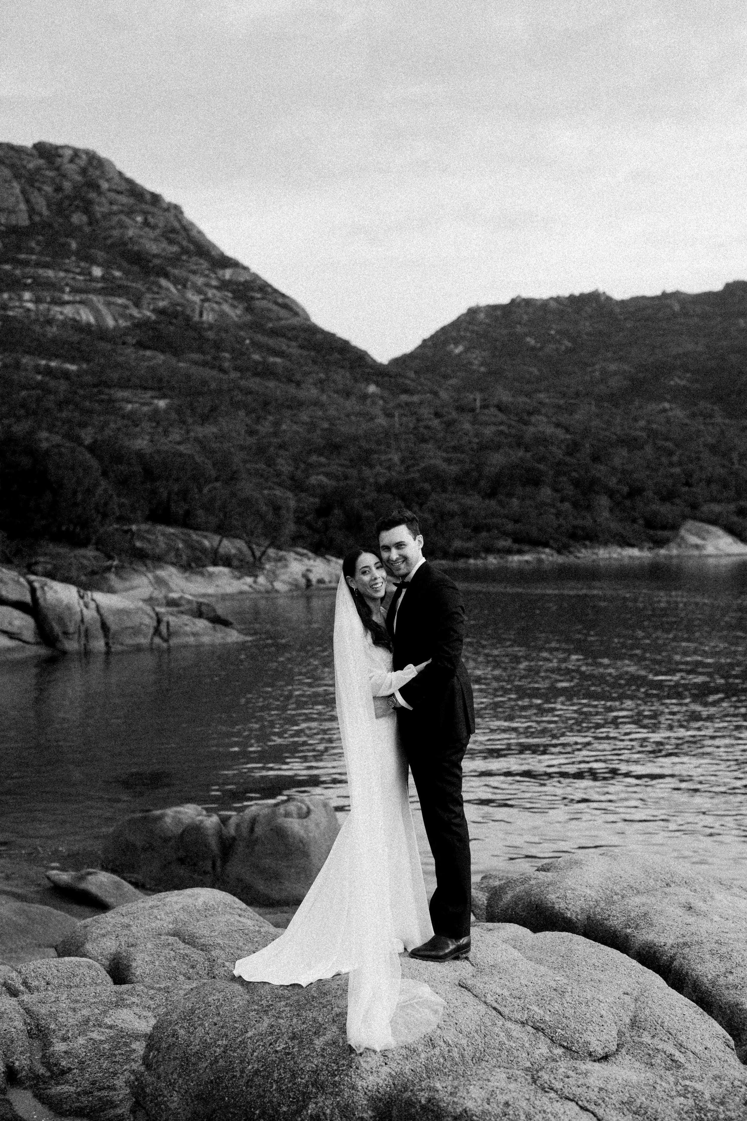 Black and white photo of a bride and groom holding each other and smiling on a rocky lakeshore with mountains in the background.