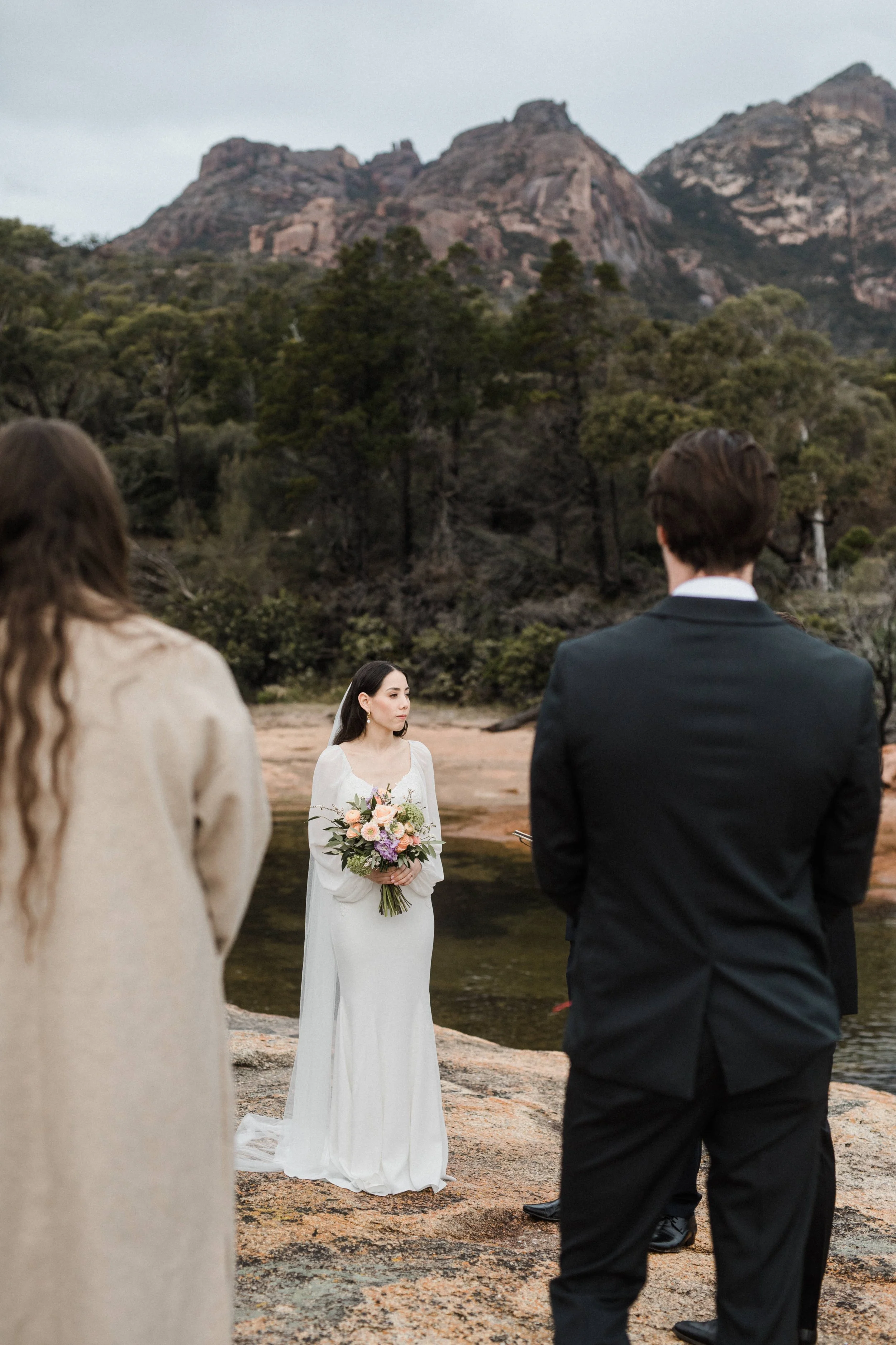 A wedding ceremony outdoors near a river with mountains in the background. The bride is holding a bouquet of flowers and standing in front of the groom and two other individuals, all dressed formally.