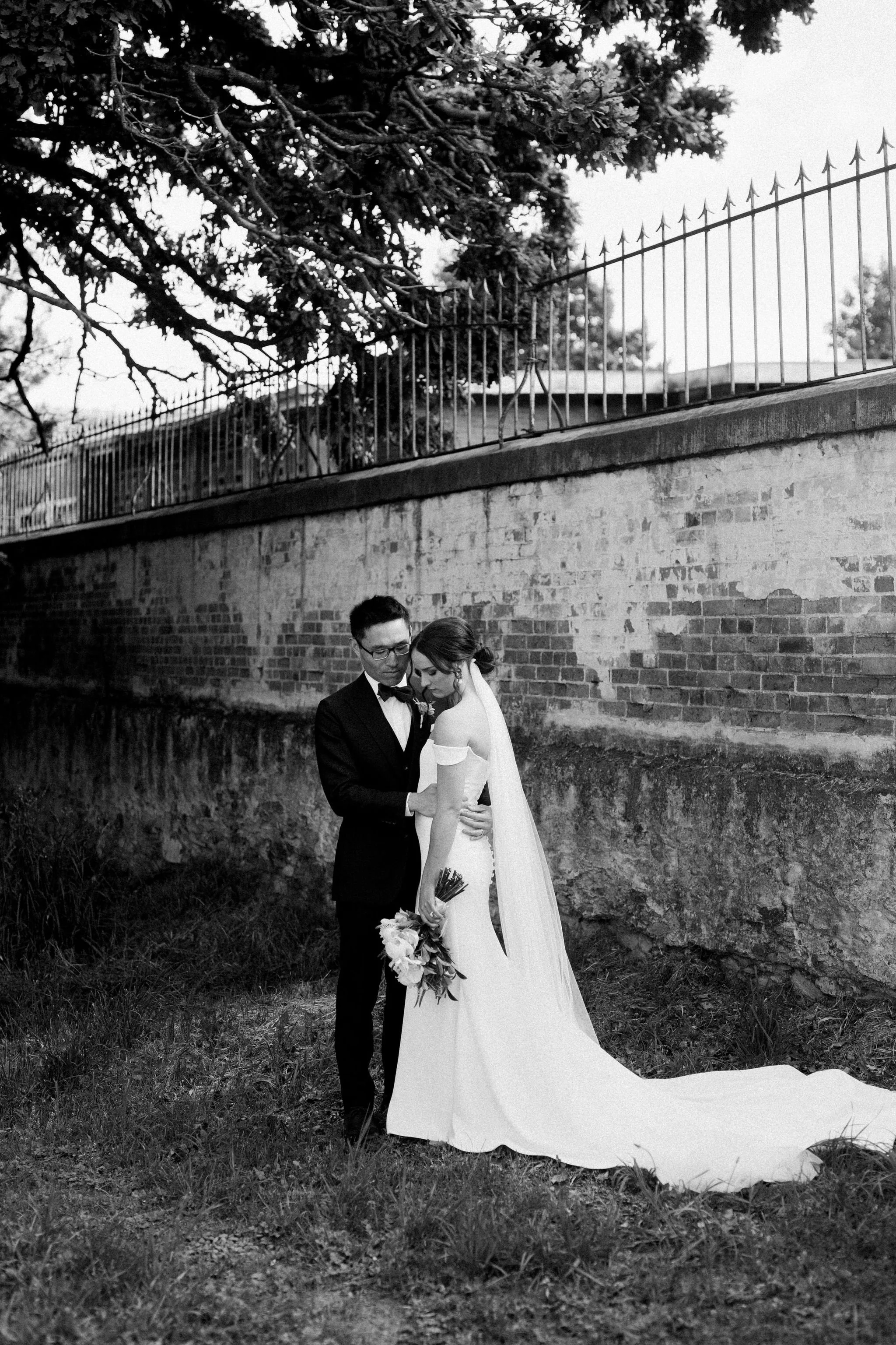 Black and white photo of a bride and groom standing closely outdoors next to a brick wall with a wrought iron fence on top. The bride is in a wedding dress holding a bouquet, and the groom is in a tuxedo. They appear to be sharing a tender moment.