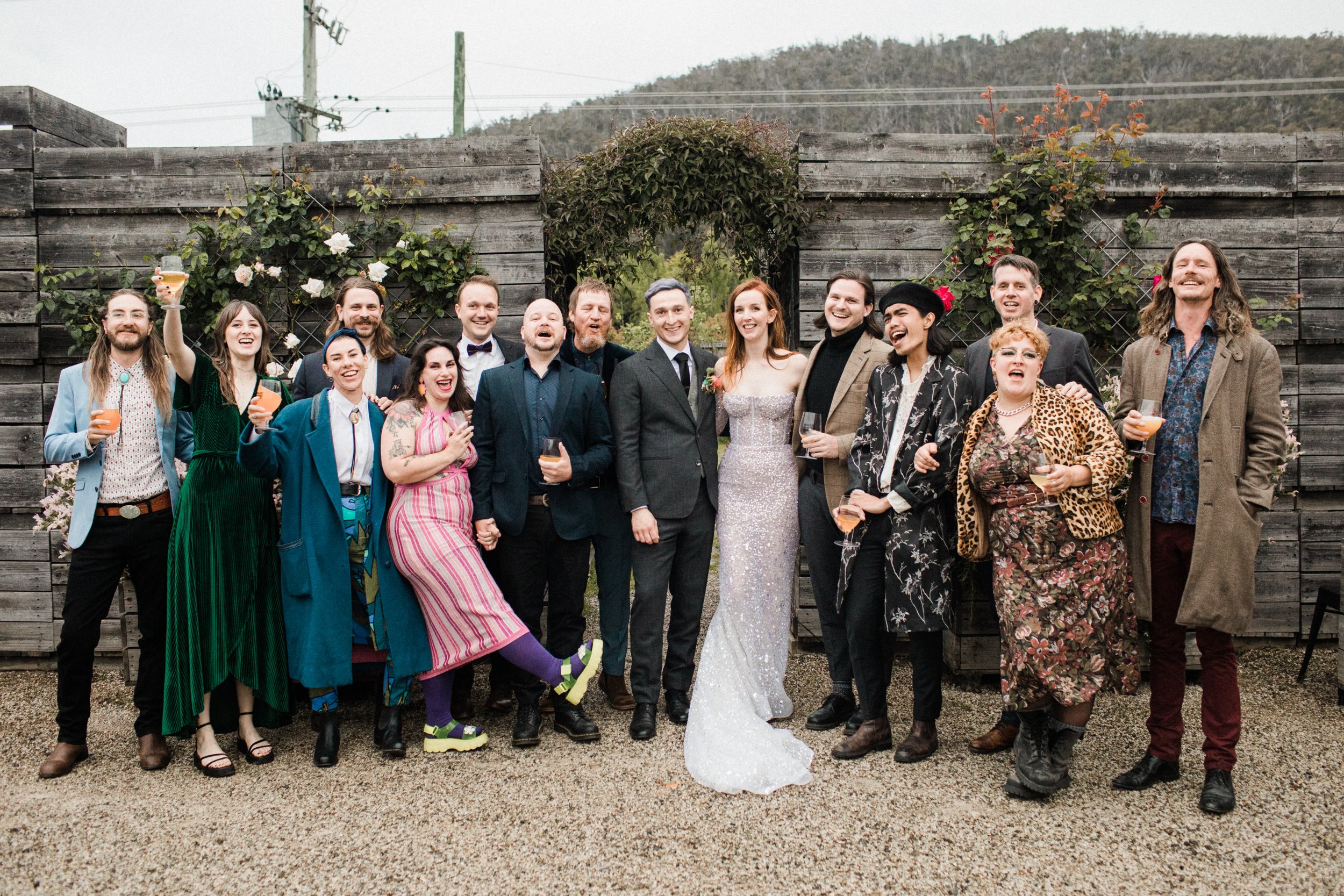 Group of people at a wedding celebration, standing outdoors in front of a wooden fence with plants, holding drinks, smiling and posing for the camera.