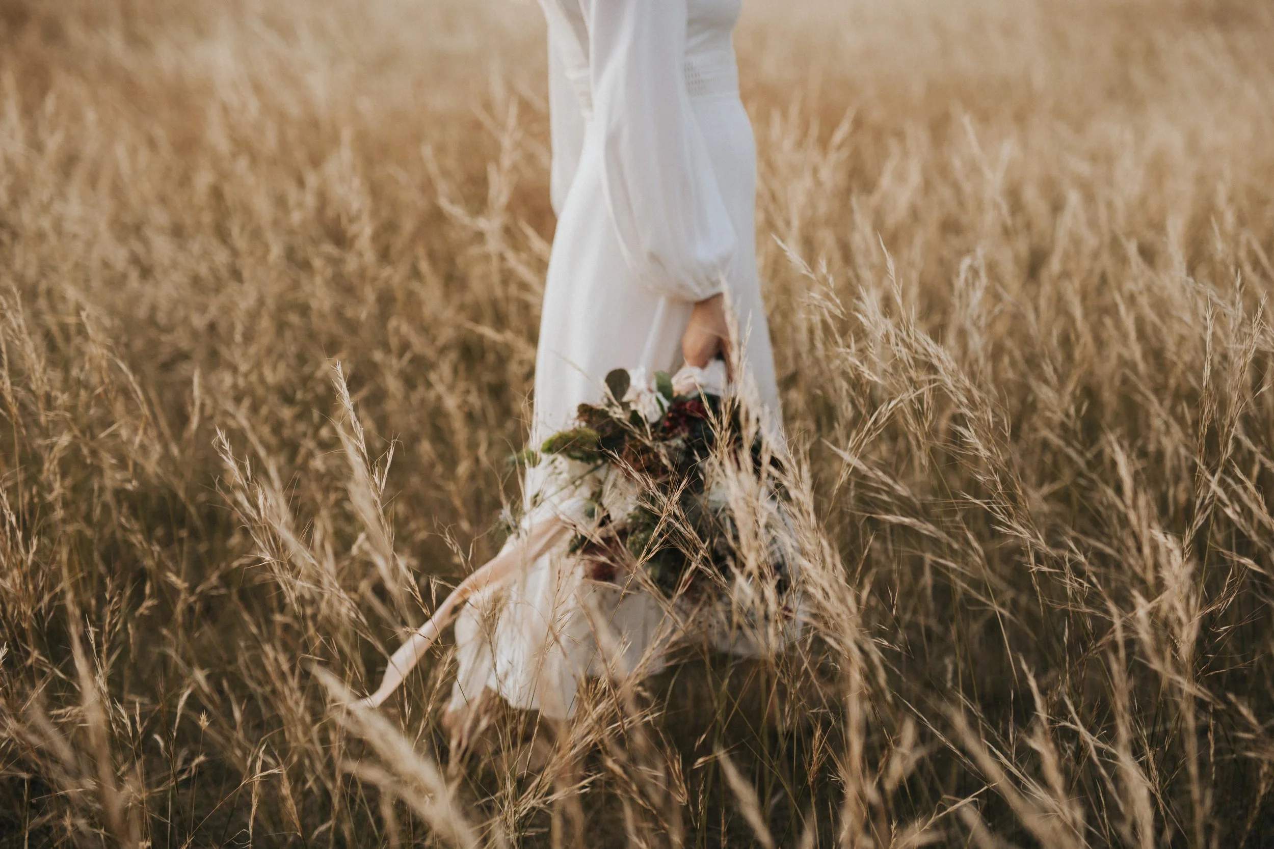 Person in a long white dress holding a bouquet of flowers walking through a field of tall, dry grass at Piermont, Tasmania.