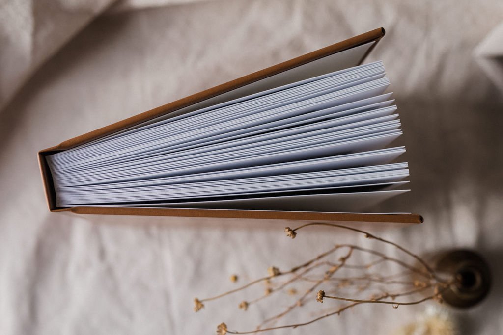 Top view of a closed hardcover book with white pages, placed on a light-colored textured surface, with dried flowers nearby.