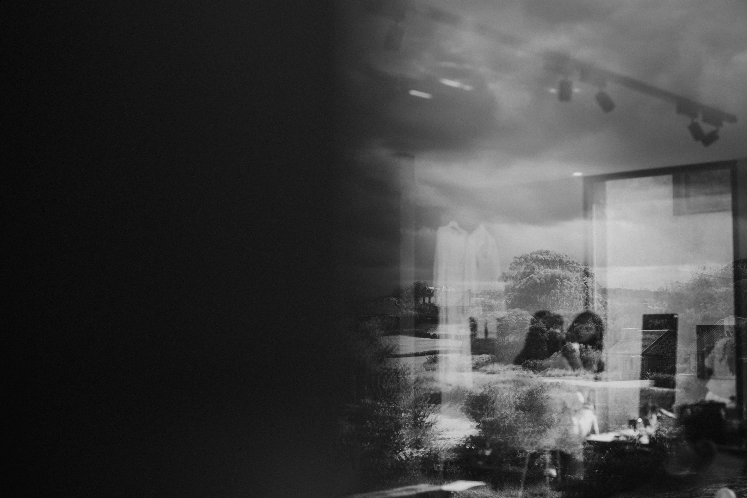 A black and white photo of a reflection in a glass window showing a park landscape with trees and a cloudy sky, indoor room with ceiling lights, a person, and furniture inside.