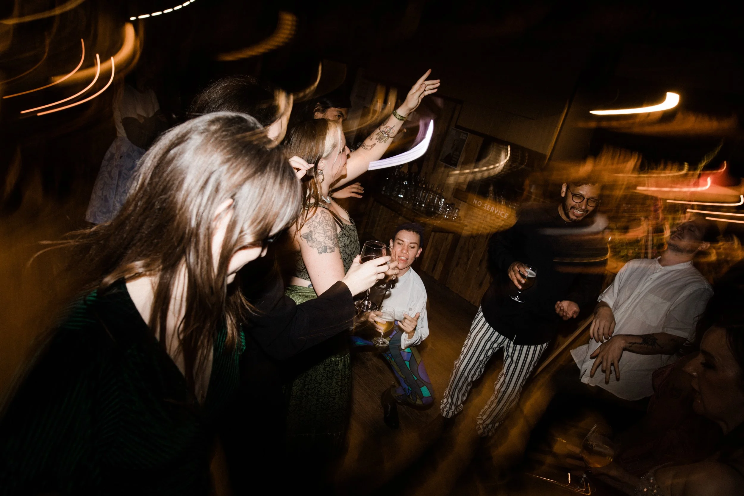 Group of people socializing and dancing in a dimly lit bar or club, holding drinks, with motion blur and light streaks.