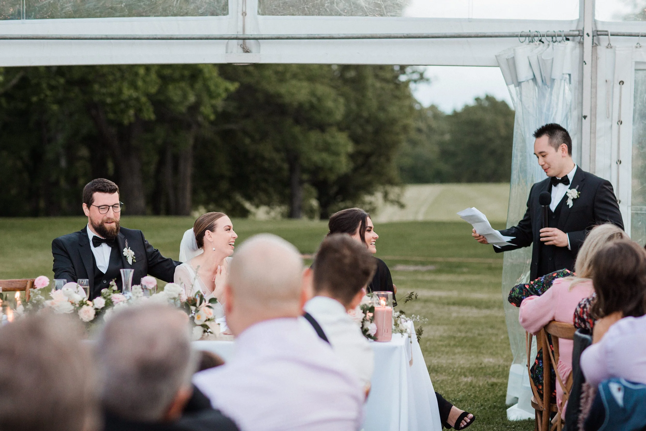 A wedding reception outdoors showing the bride and groom sitting at a table, smiling and listening to a man giving a speech with a microphone, while guests seated around watch and listen in a grassy area with trees in the background.