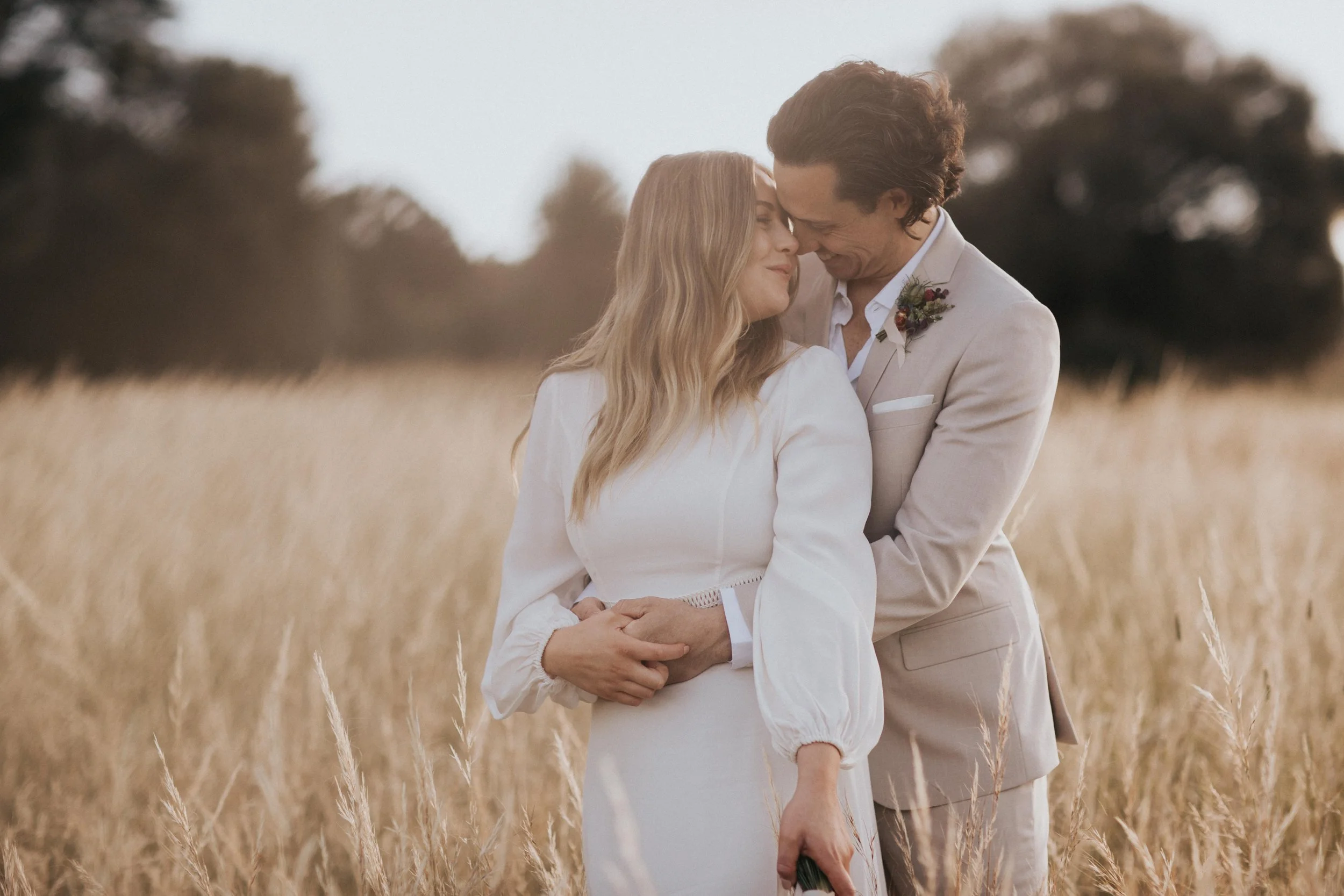 A couple dressed in wedding attire embracing in a field of tall, golden grass with trees in the background during sunset or late afternoon.