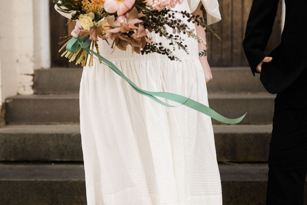 Person in a white dress holding a bouquet of flowers with pink, cream, and purple blooms, and a light green ribbon, standing near another person in a dark suit.