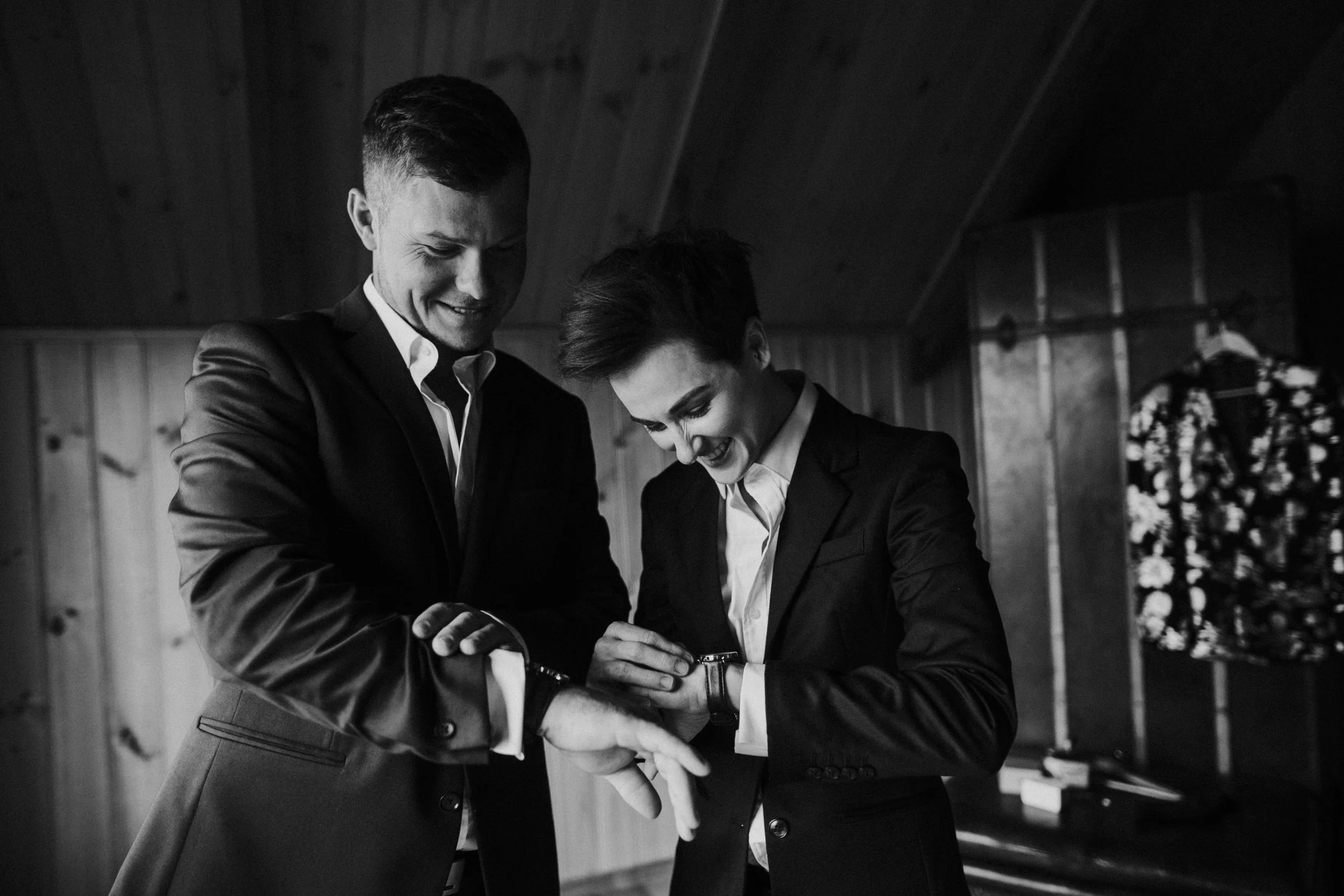 Two people in formal suits smiling and laughing in a wooden room, with one checking the other's watch.