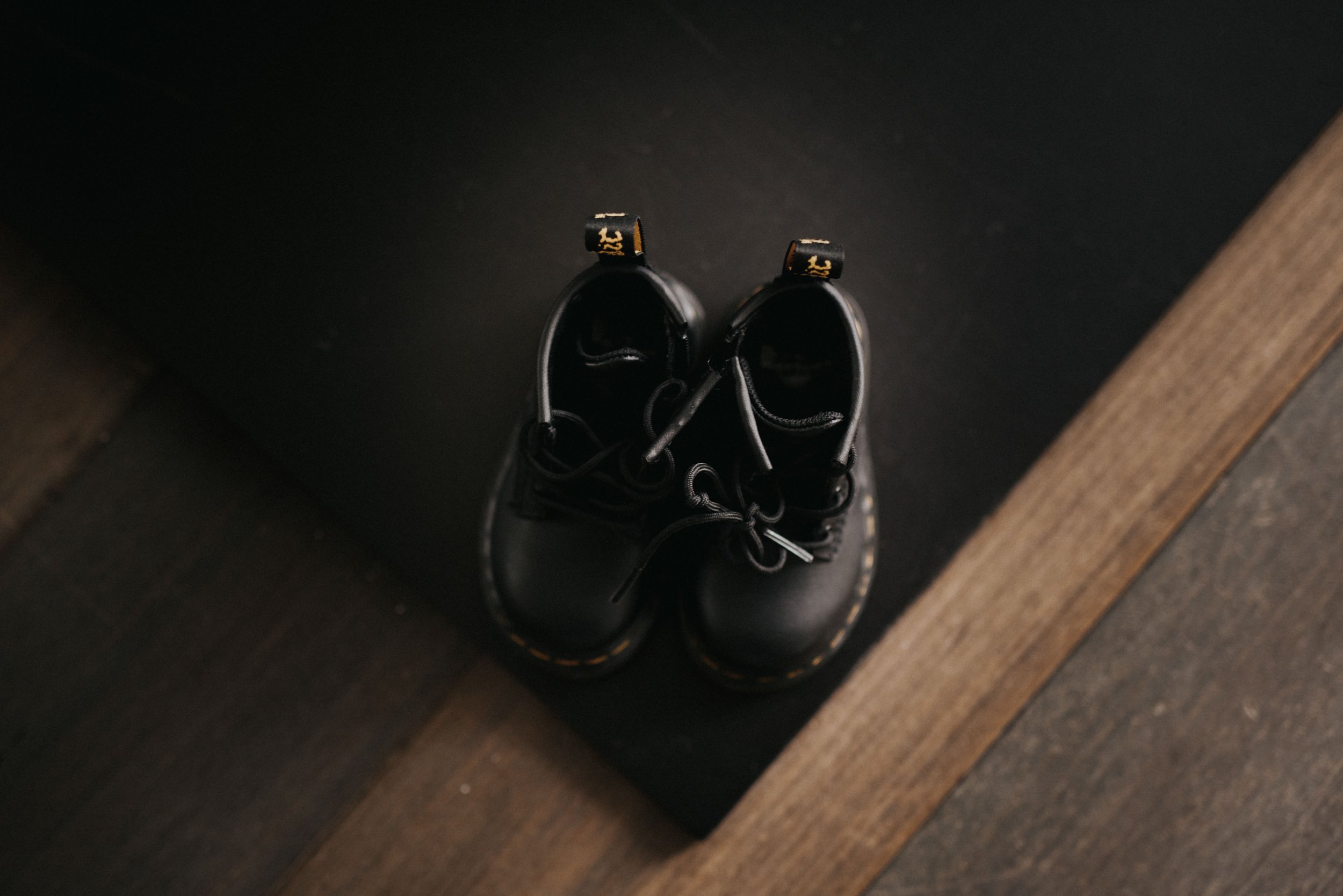 Pair of black boots with yellow stitching, viewed from above on a dark mat next to a wooden floor.