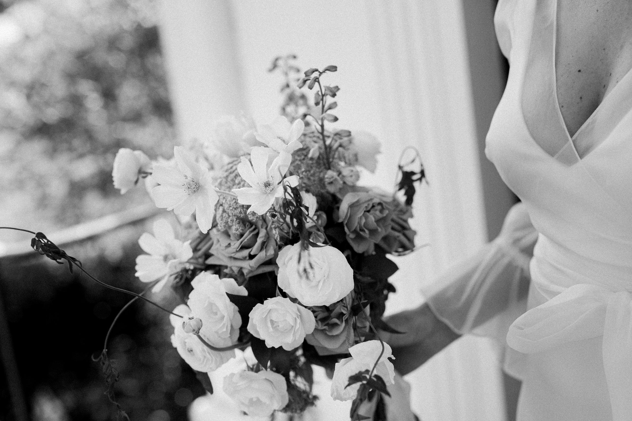 Close-up of a bride holding a bouquet of flowers, including roses and other blossoms, in black-and-white.
