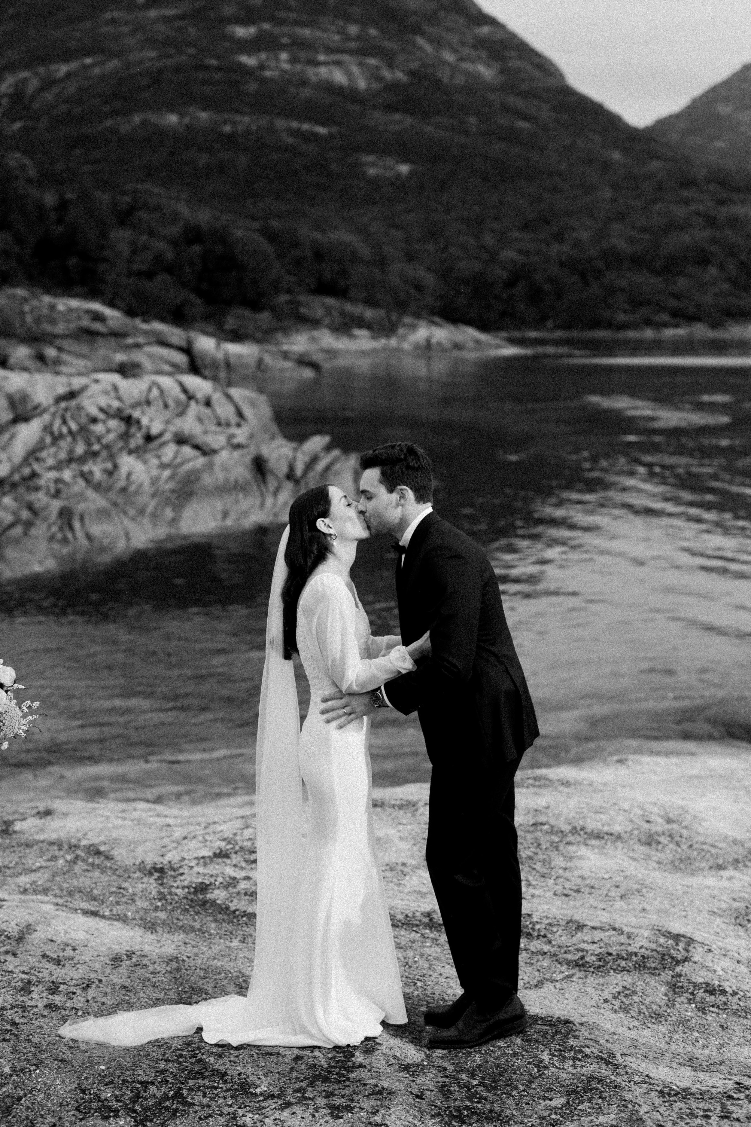 A black-and-white photograph of a bride and groom kissing by a lake with mountains in the background.