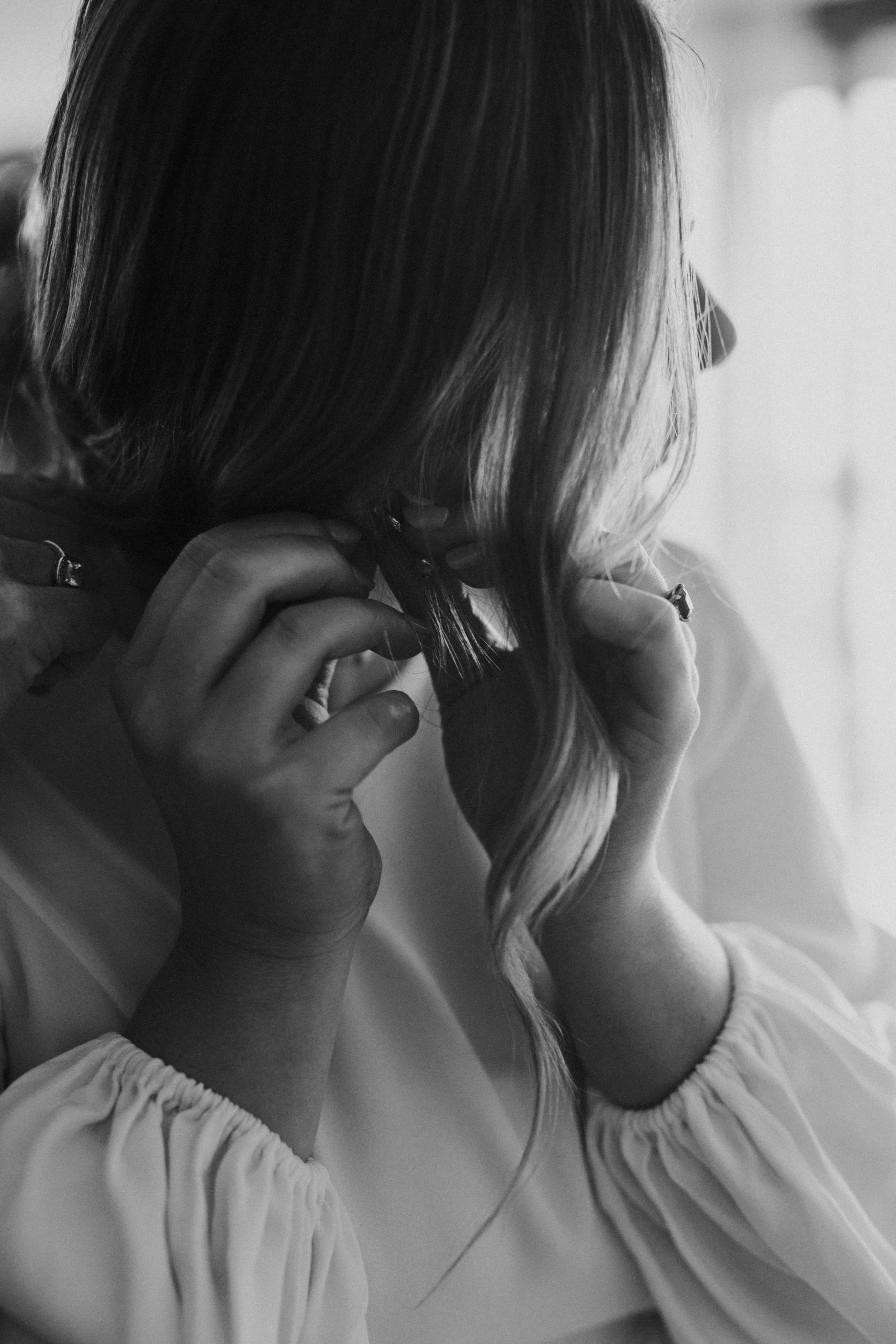A woman with long, wavy hair, wearing a light-colored top with gathered sleeves, is touching her hair with her left hand and adjusting it with her right hand.