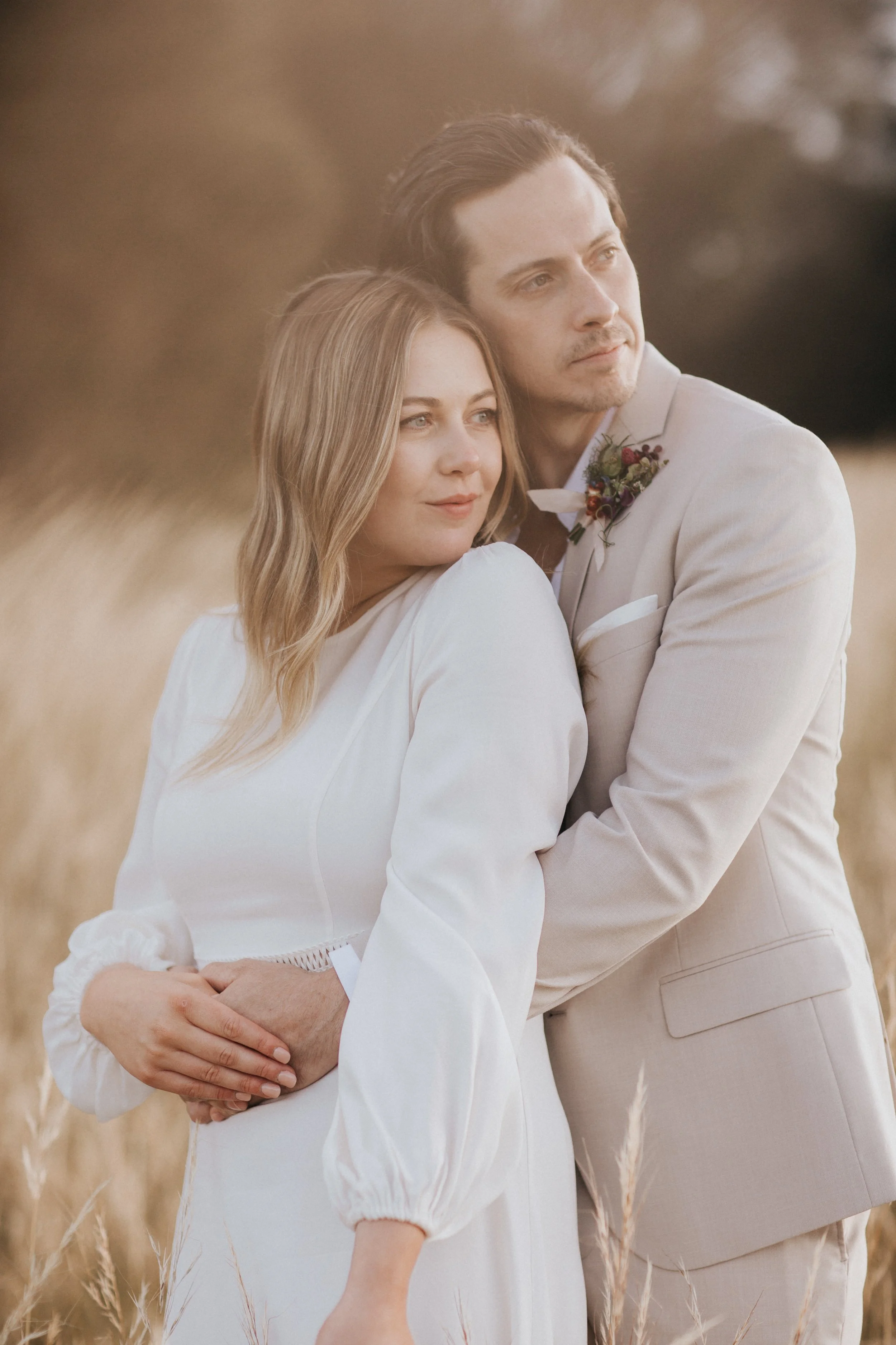 A couple embraces in a golden field during sunset, dressed in wedding attire, holding each other tenderly with a blurred background of trees and tall grass.