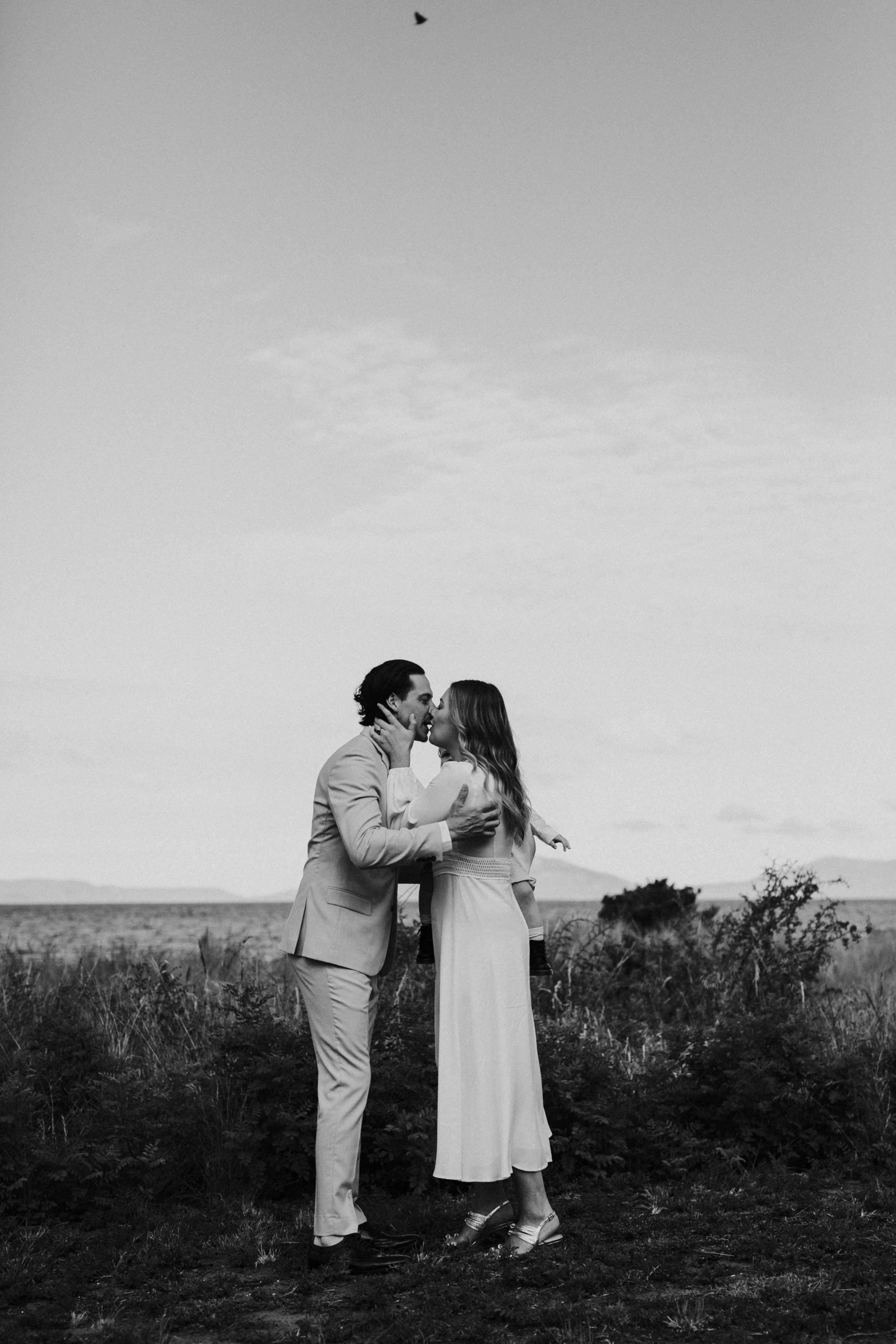 A black and white photo of a couple kissing outdoors with a child on the woman's hip, in a grassy field with bushes and distant mountains, and a bird flying in the sky.