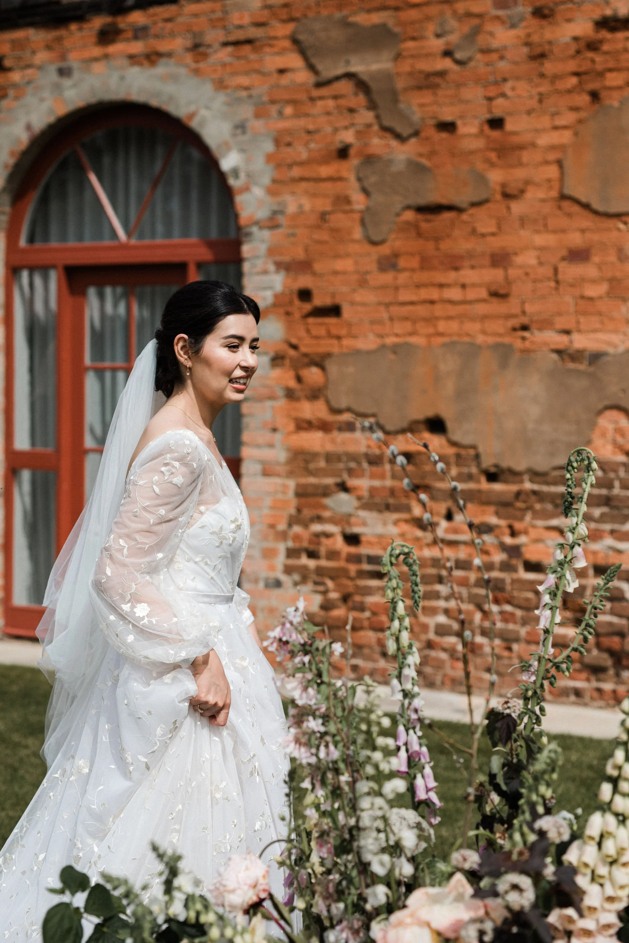 A bride in a white wedding gown with floral embroidery, standing outdoors near a brick wall and arched window, smiling and looking to the side, with flowers in the foreground.
