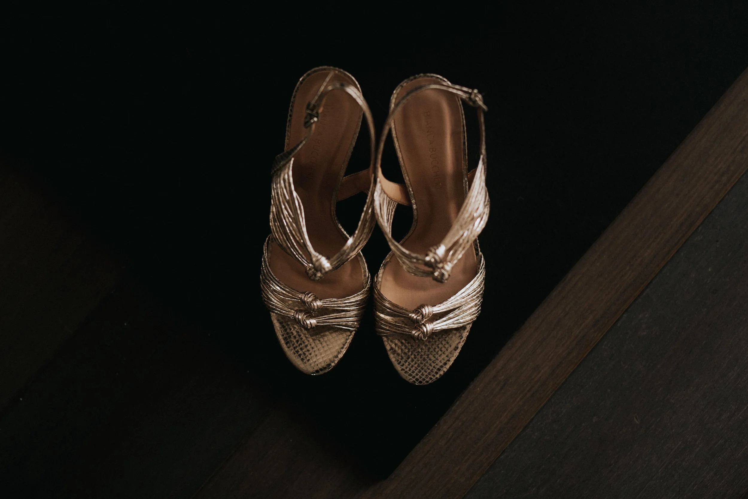 A pair of metallic high-heeled sandals with intricate strap detailing, viewed from above on a dark surface.