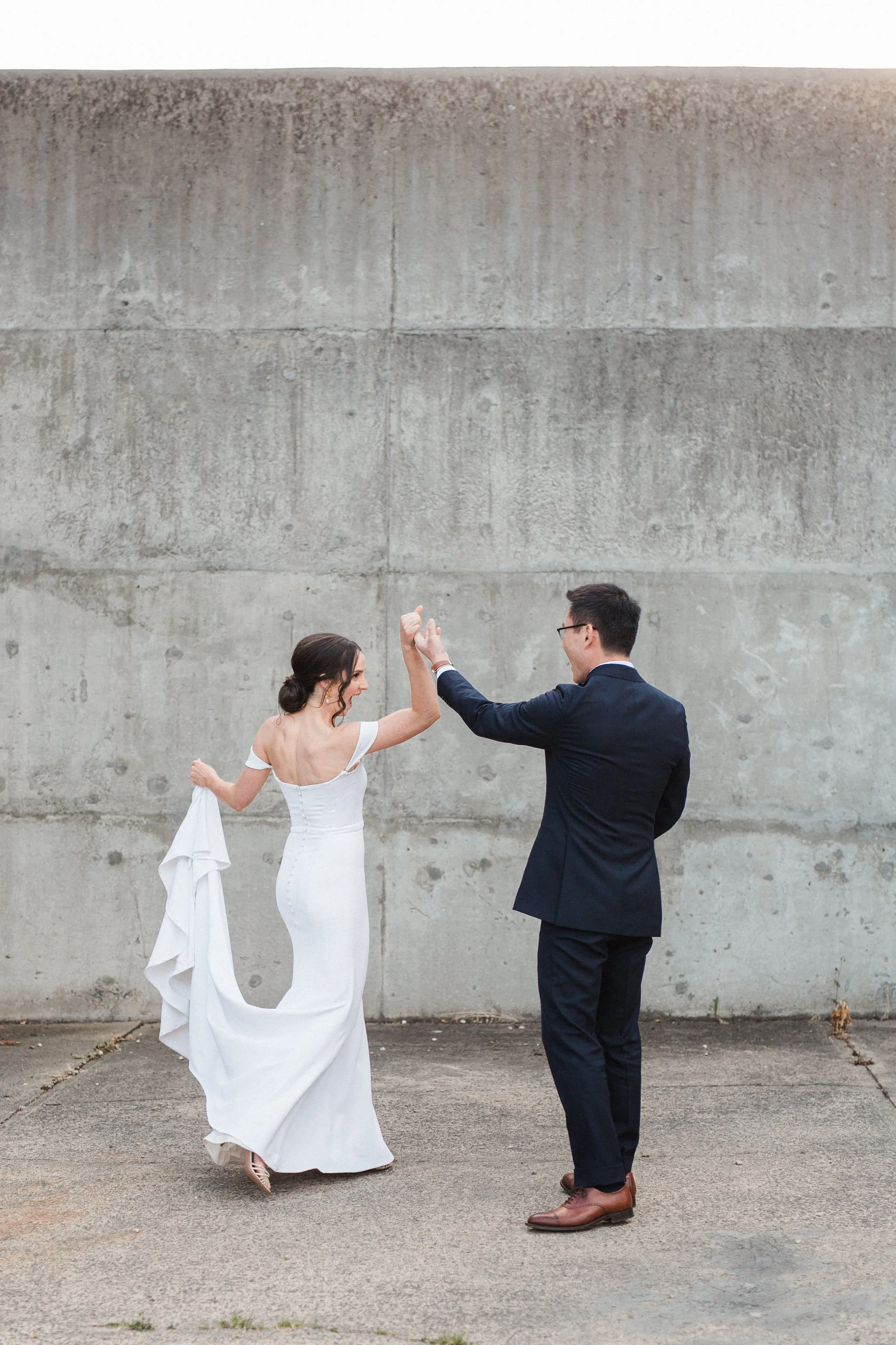 A bride and groom dancing outdoors in front of a plain concrete wall.