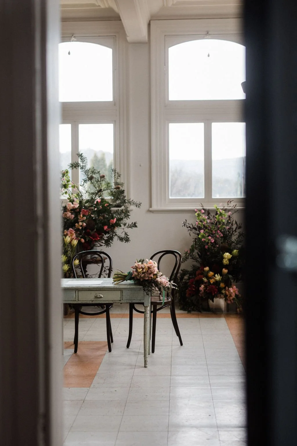 A room with large windows, a light-colored wooden table with a flower bouquet, two black chairs, and numerous colorful flowers in the background.
