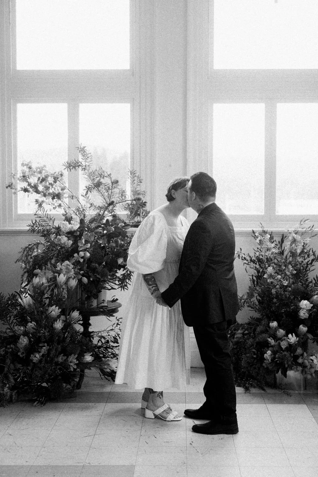 A couple sharing a kiss during a wedding ceremony, standing in front of large windows and floral arrangements.
